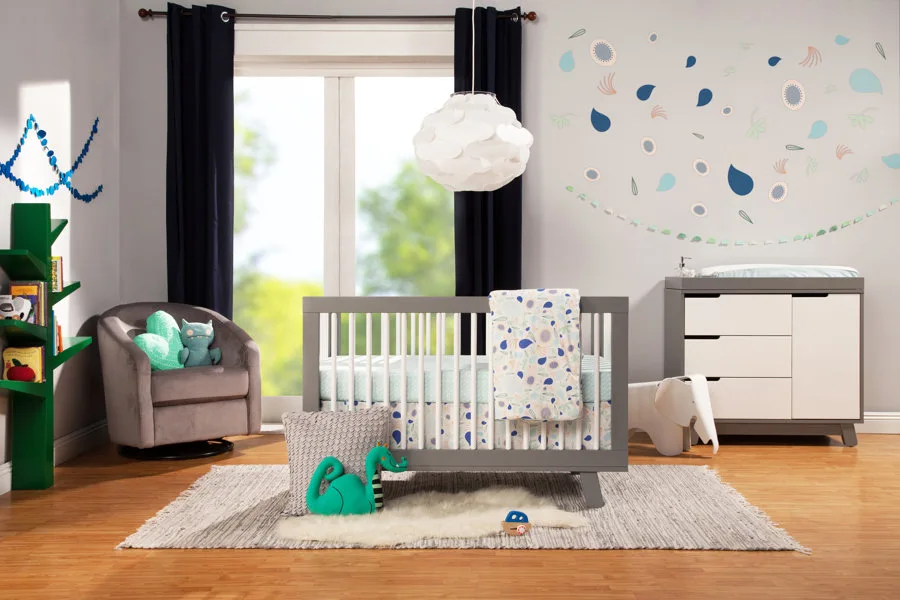 A modern nursery features a gray wooden crib, a matching changing table, a green tree-shaped bookshelf, and colorful wall decals on a gray wall.