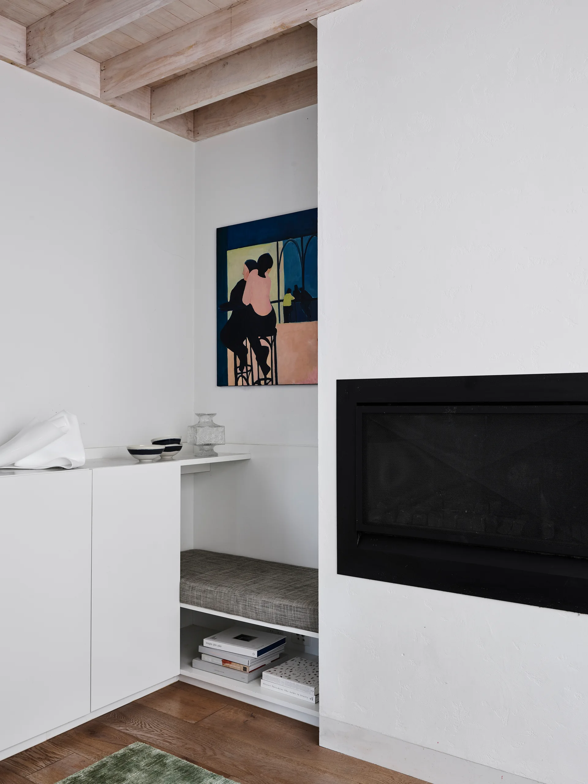 A black fireplace is integrated into a textured white plaster wall next to a white cabinet with an inset grey bench cushion.