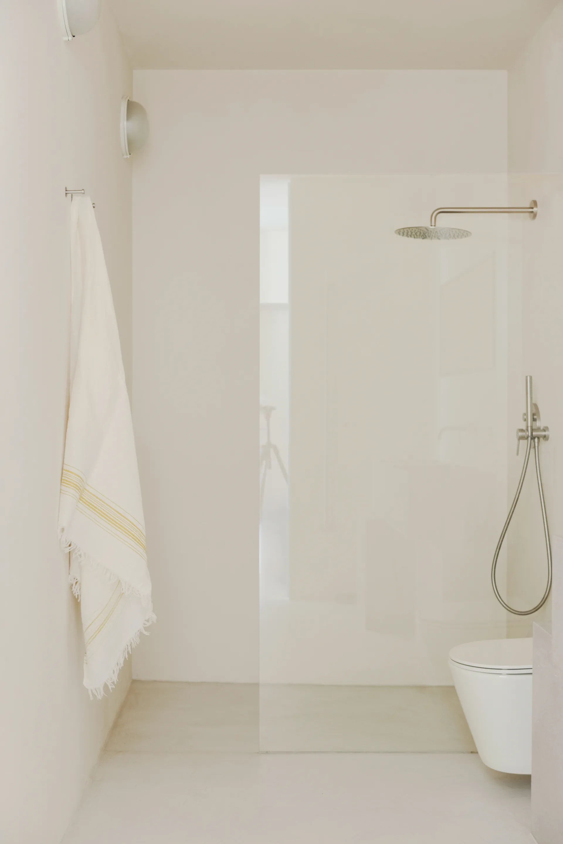 A minimalist off-white bathroom featuring a walk-in shower with a frameless glass screen, a wall-mounted toilet, and a fringed towel.