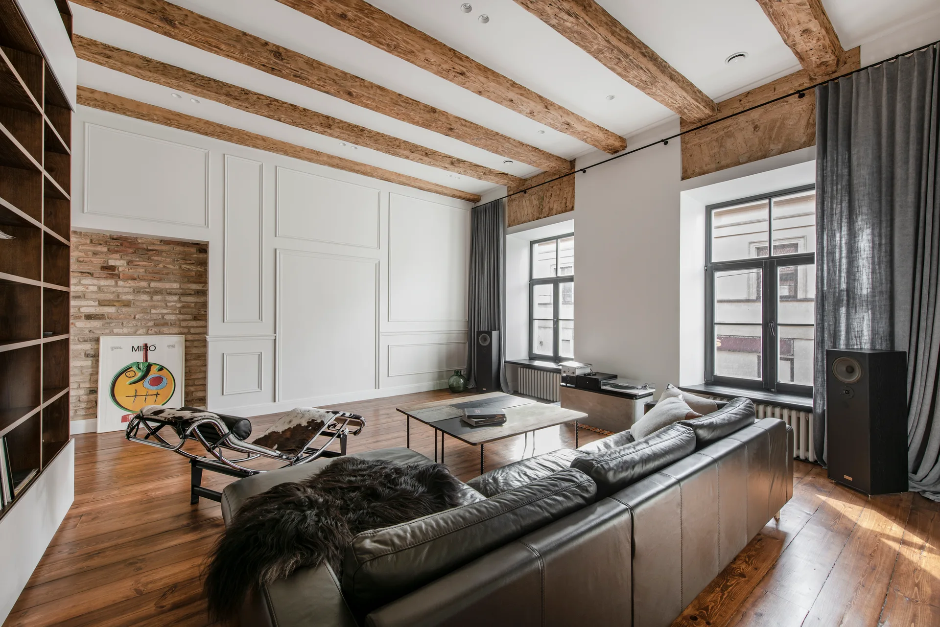 A spacious living room features a dark leather sofa, exposed wooden ceiling beams, white paneled walls, and large windows with grey drapes.