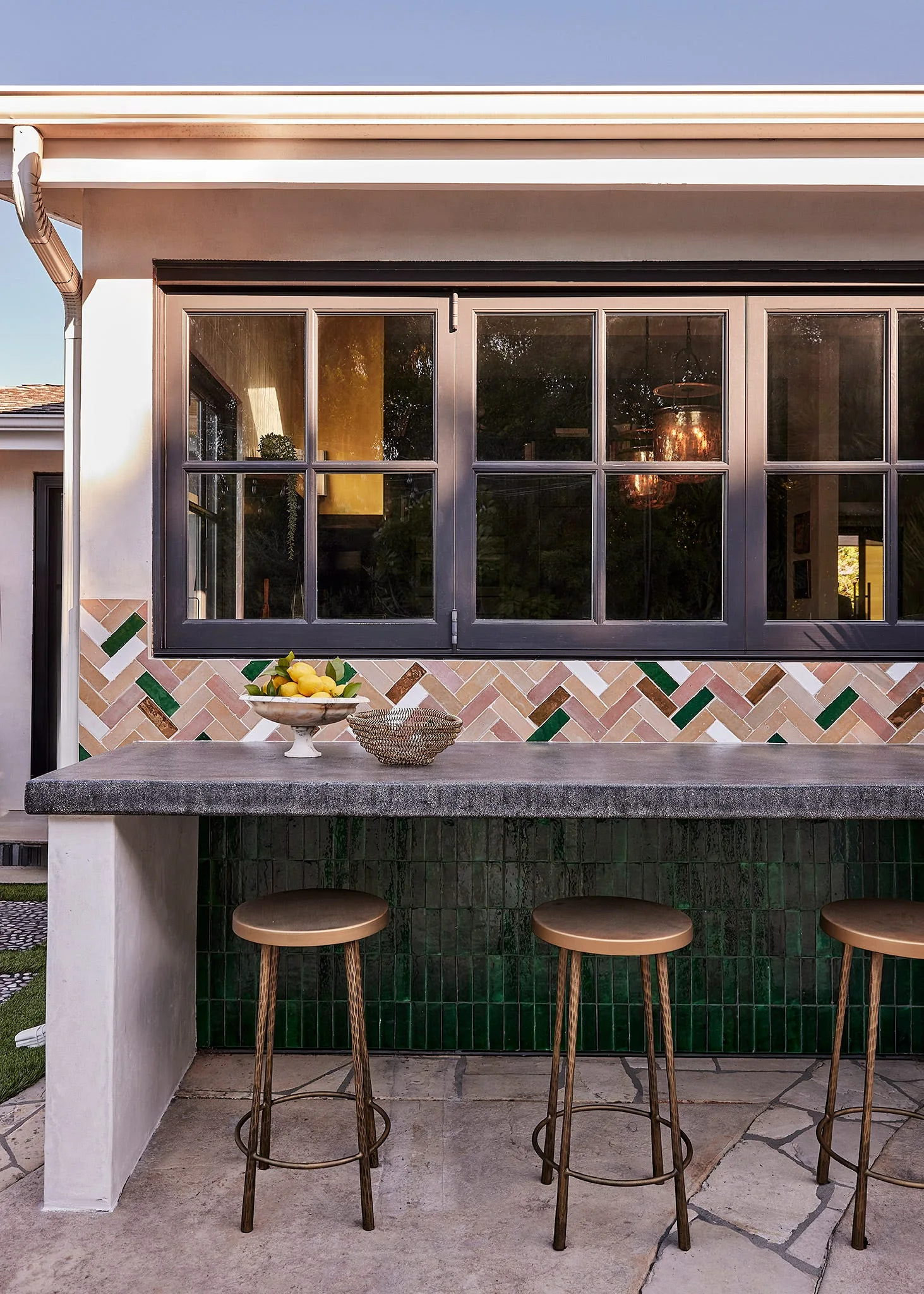 An outdoor stone bar counter features gold metal stools, a green-tiled base, and a multi-colored herringbone tile backsplash under a window.