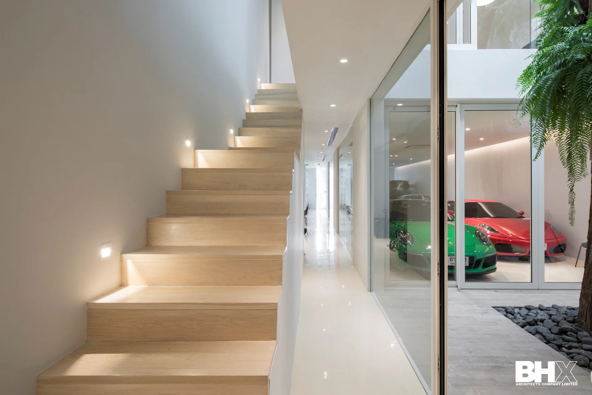 A minimalist hallway features a light wood staircase with recessed wall lights and a glass partition looking into a garage with cars.