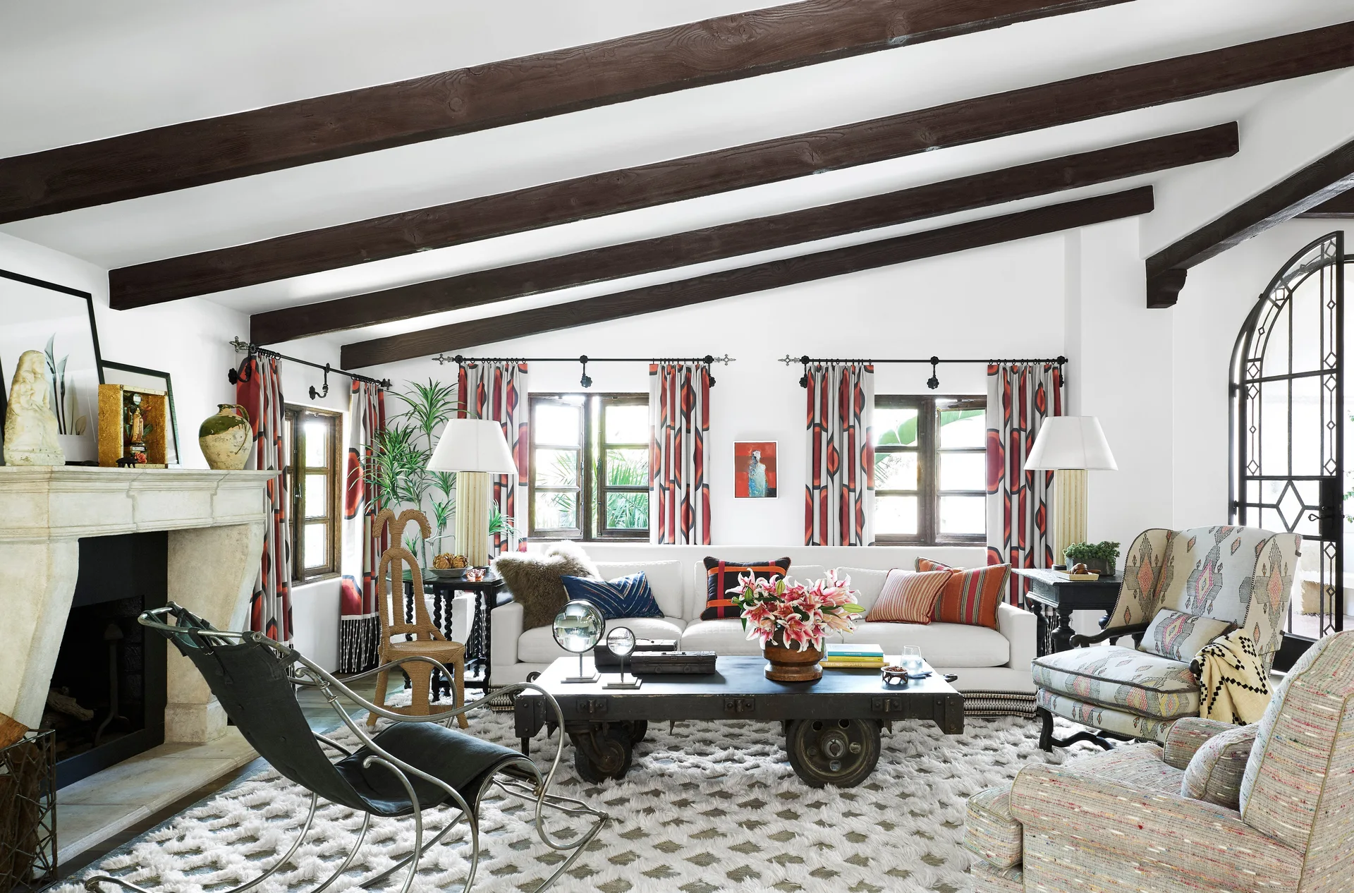 An eclectic living room with dark exposed ceiling beams, a white sofa, a wheeled coffee table, and red patterned curtains framing three windows.