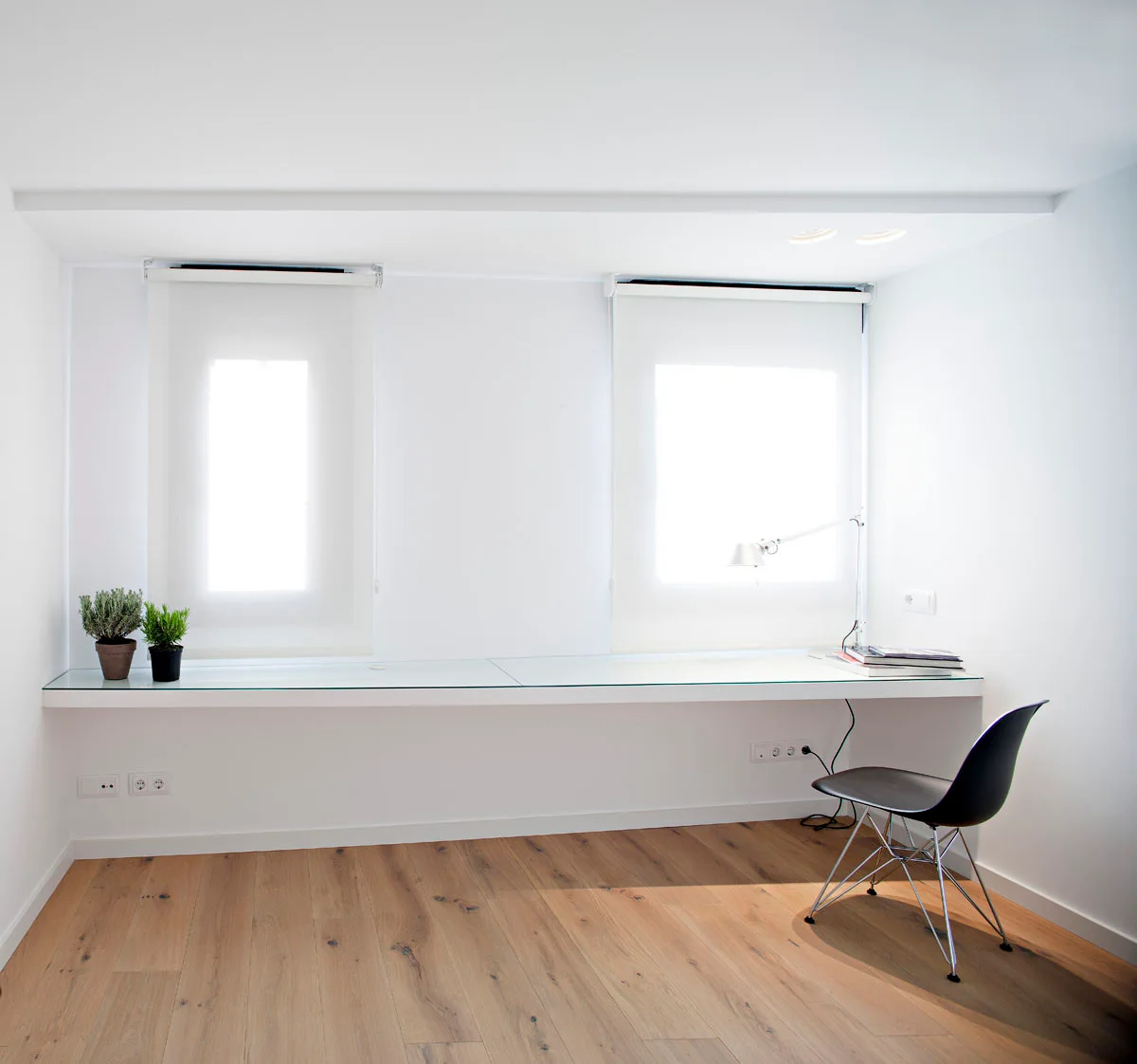 A long floating white desk with a glass top is paired with a black chair in a minimalist white office with wood flooring.