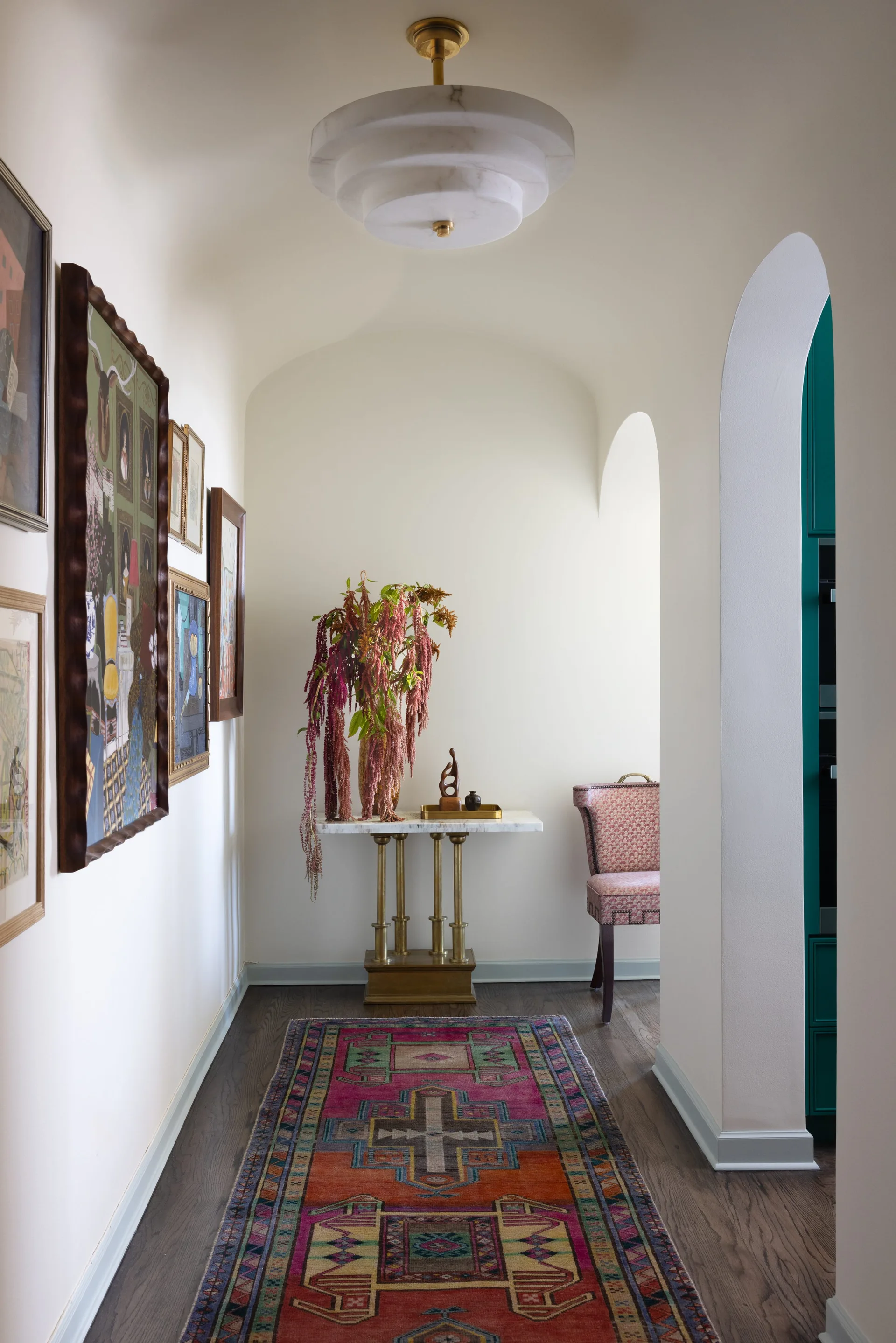 A cream hallway features a colorful patterned runner rug, a gallery wall of framed art, and a marble-top console with draping flowers.