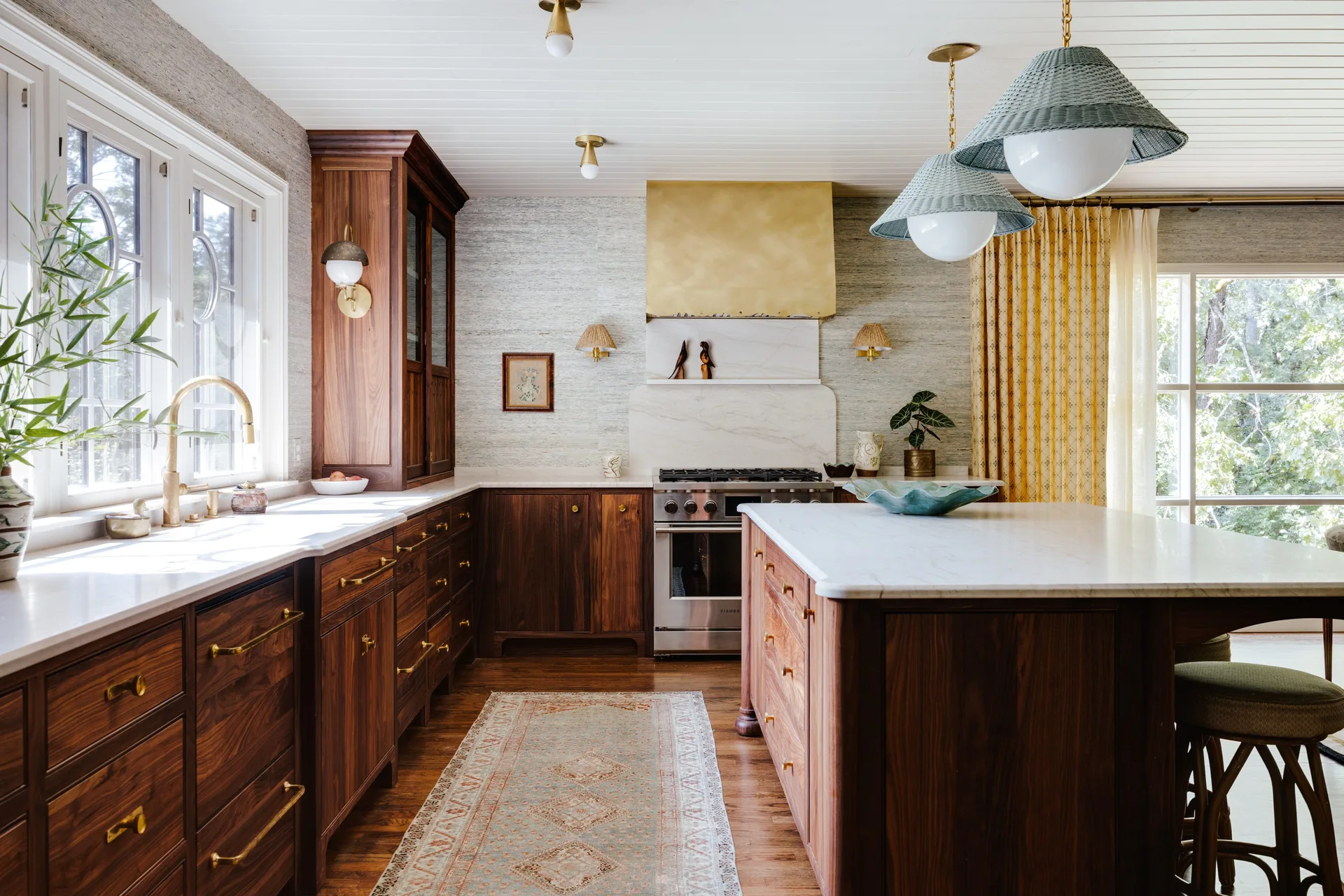 A kitchen featuring dark wood cabinetry, a marble-topped island, a brass range hood, and textured wallpaper with two green pendant lights.