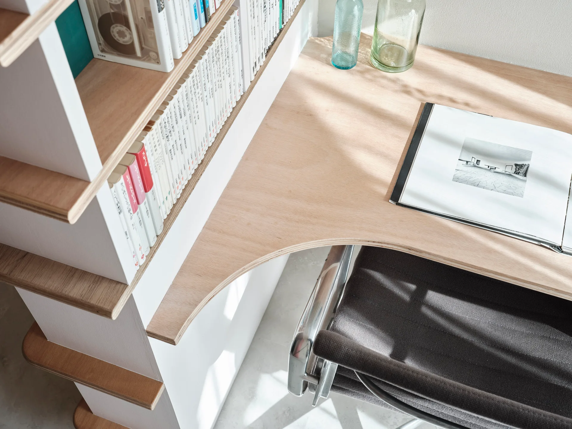 An integrated light wood desk with a curved inner edge and exposed plywood trim meets a white bookshelf filled with books next to a dark chair.