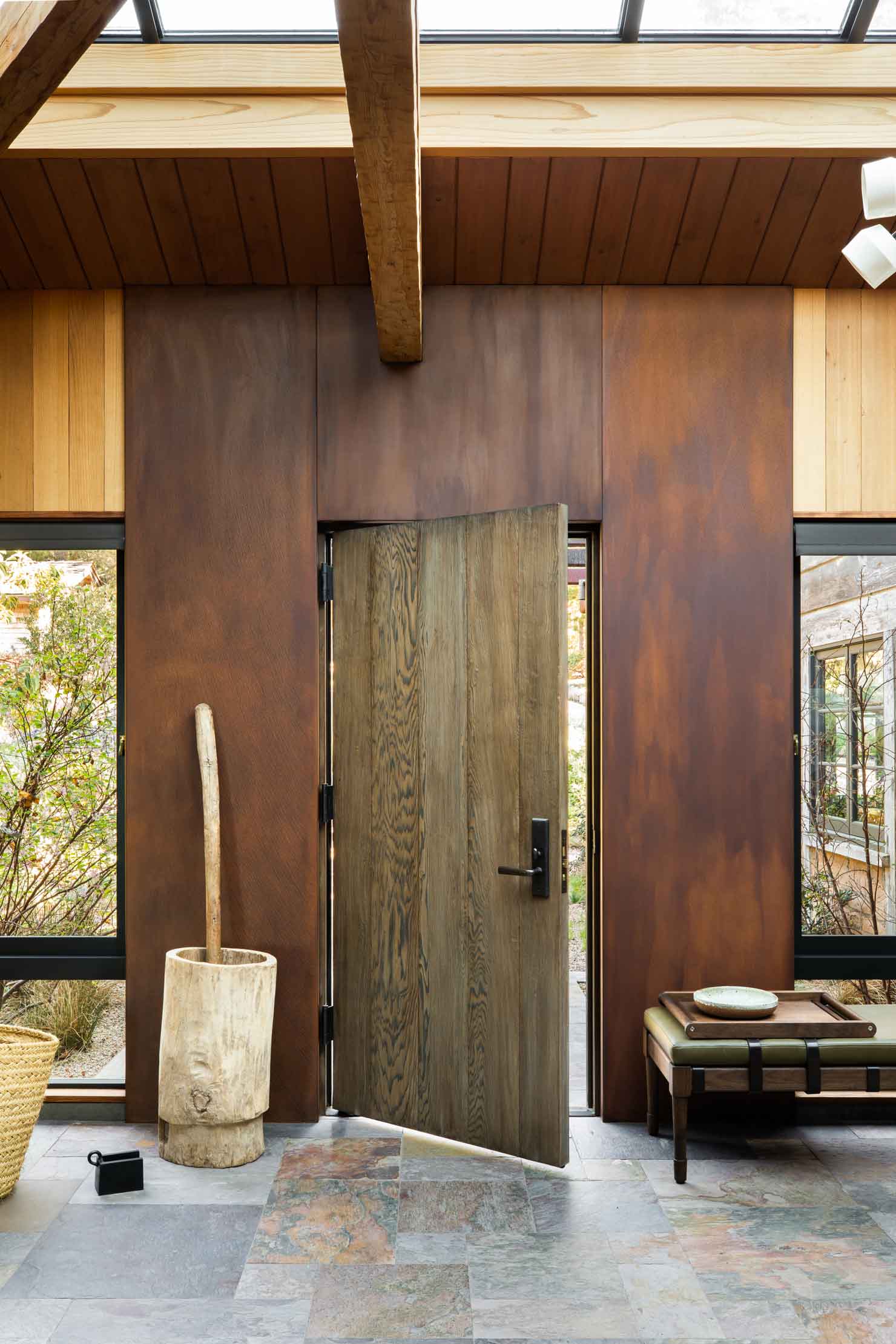 A rustic entryway features an open wooden door flanked by rusted metal wall panels, slate floor tiles, and a beamed ceiling with skylights.