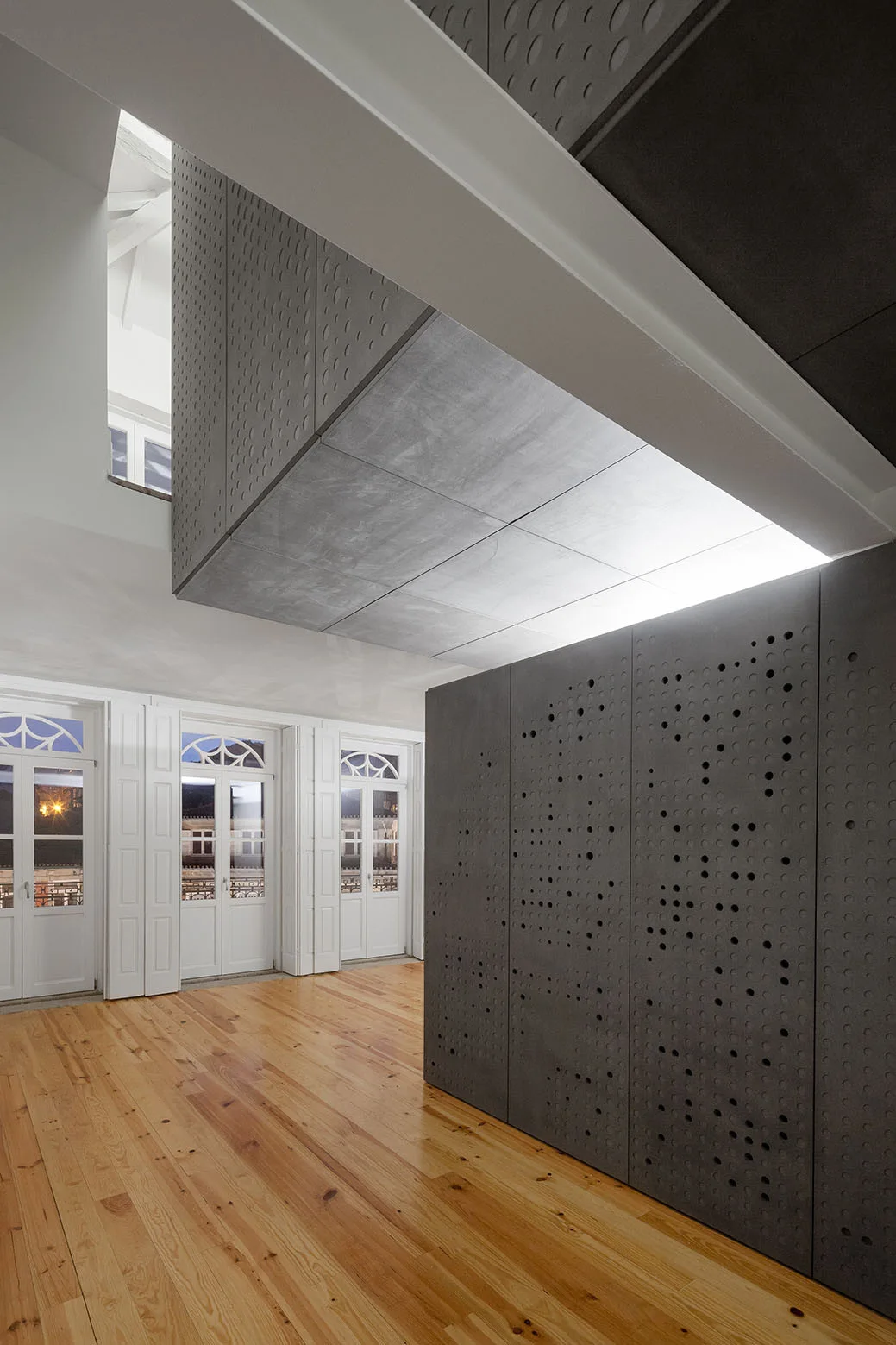 A dark grey perforated partition wall stands on glossy wood floors next to historic white arched windows in a minimalist room.