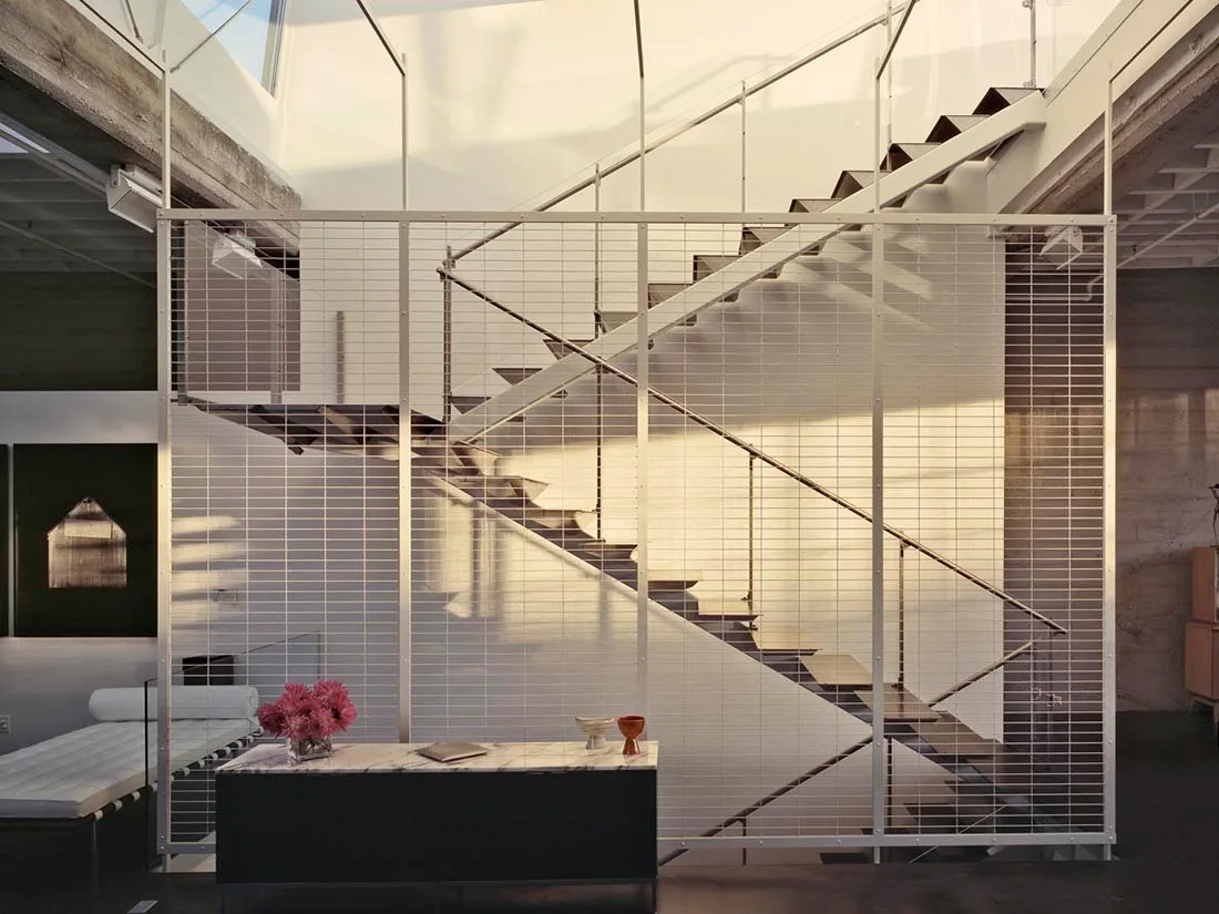 An industrial-style metal staircase is visible behind a large white metal wire mesh partition screen next to a low marble console table.