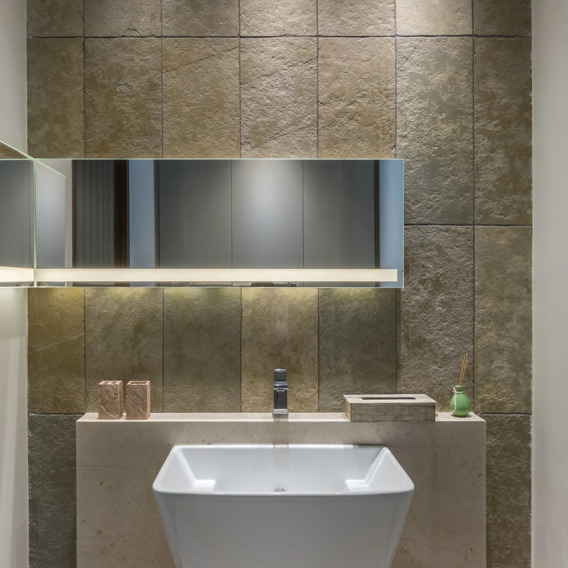 A modern bathroom vanity with a white ceramic sink and smooth beige countertop set against a rough textured stone tile wall and an illuminated mirror.