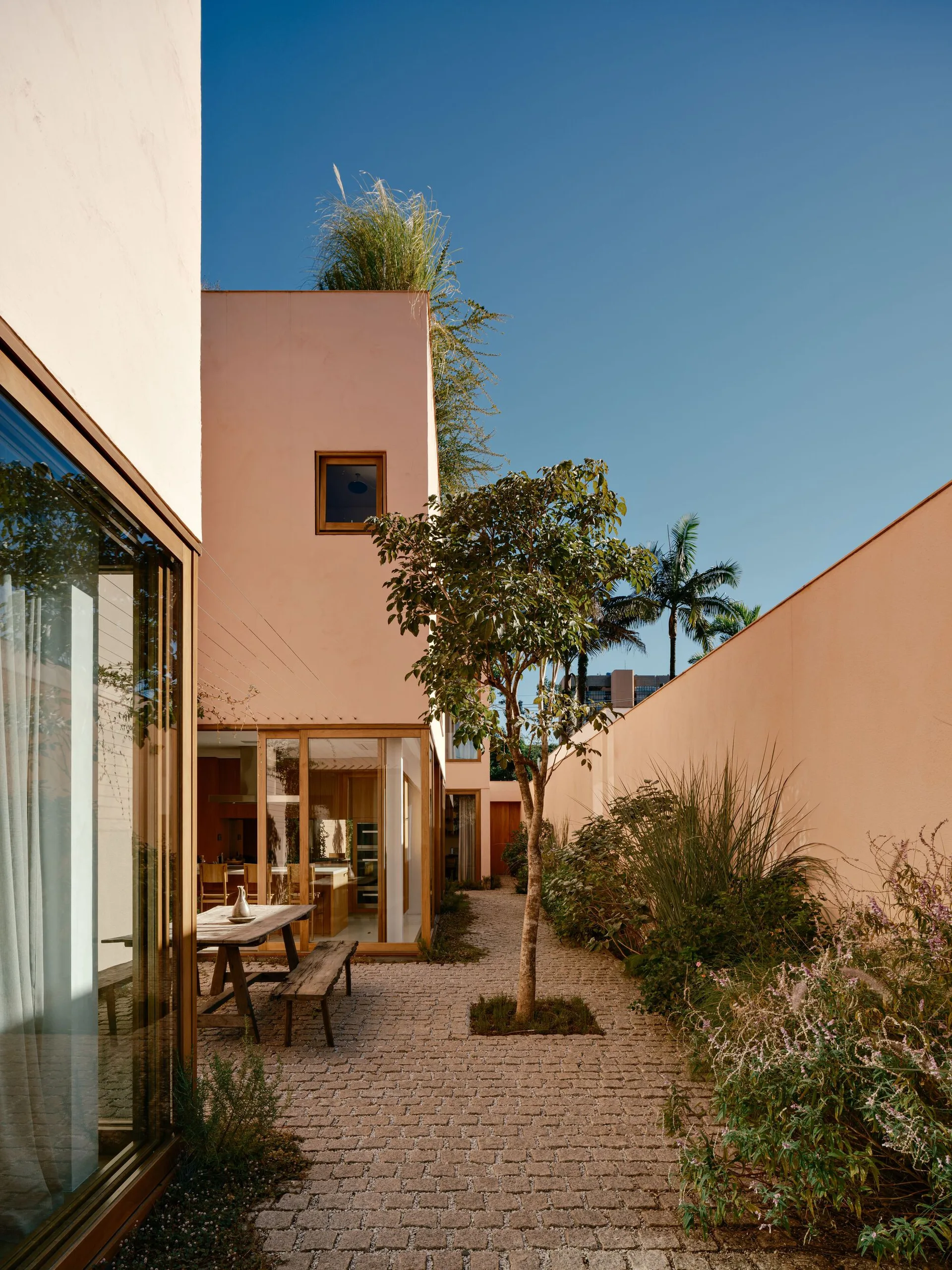 A modern outdoor courtyard featuring a wooden dining table and bench on cobblestone paving next to a pink stucco building with large windows.