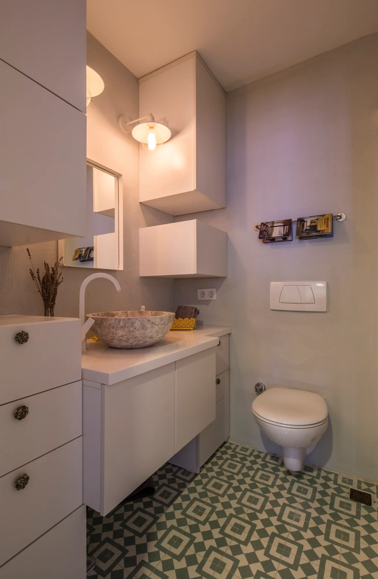 A small powder room features a white vanity with a stone vessel sink, a wall-mounted toilet, and green-and-white geometric floor tiles.