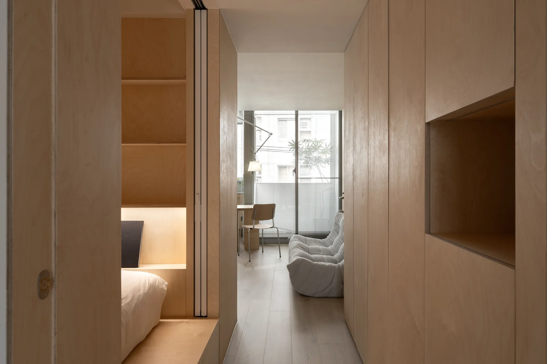 A hallway lined with custom light wood cabinetry leads from a built-in bed nook to a bright living room with a grey sofa and a window.