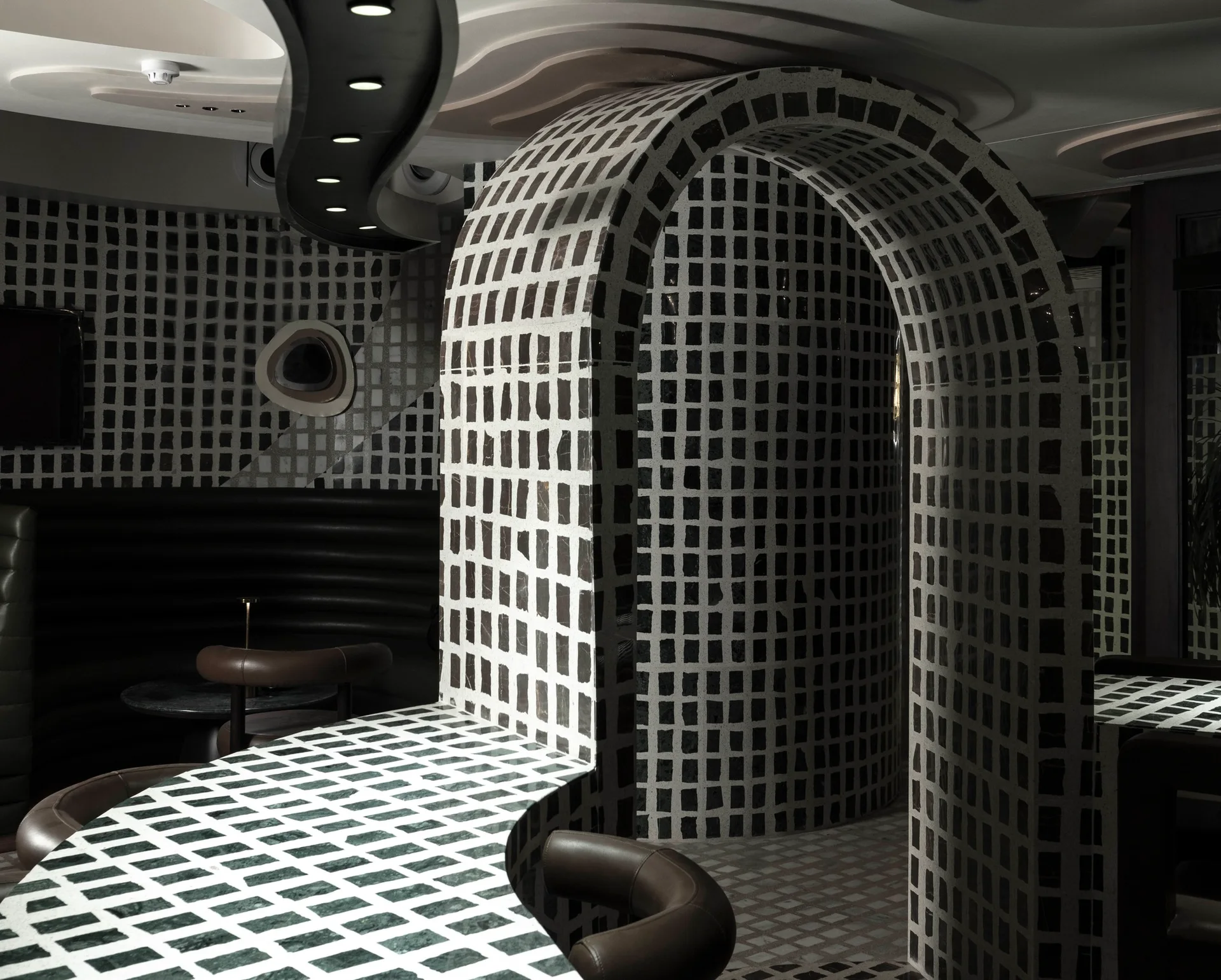 A moody interior room featuring a curved architectural arch and counter covered in a black and white grid tile pattern next to dark leather chairs.