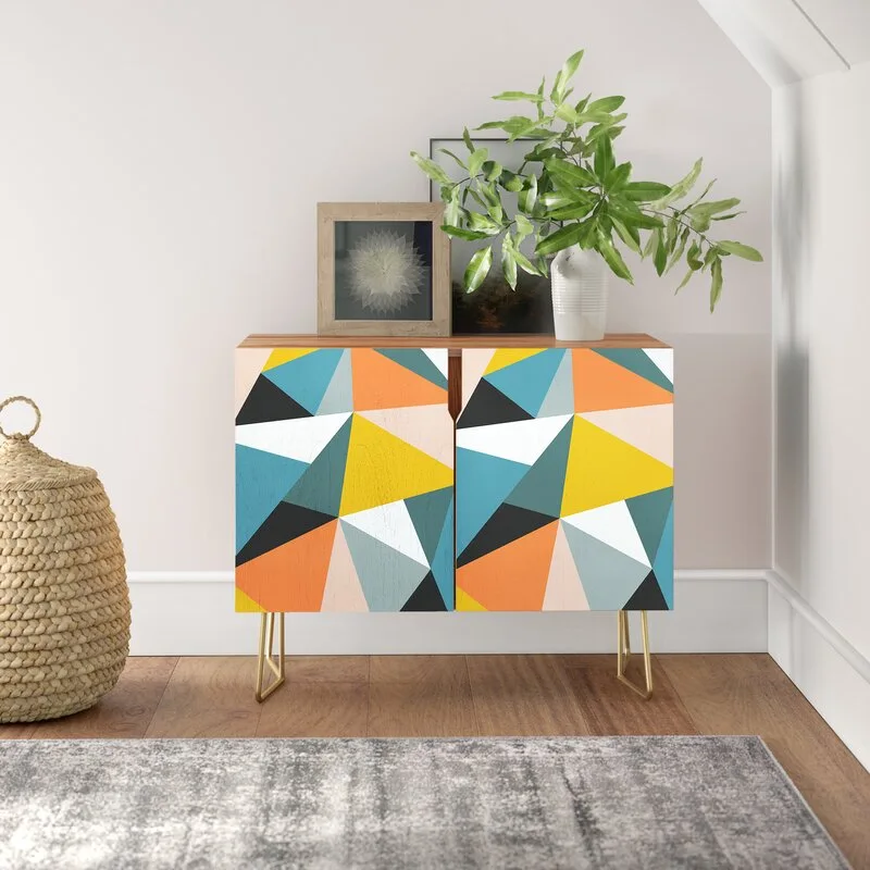 A colorful geometric patterned storage cabinet with gold hairpin legs stands on a hardwood floor next to a woven basket and grey rug.