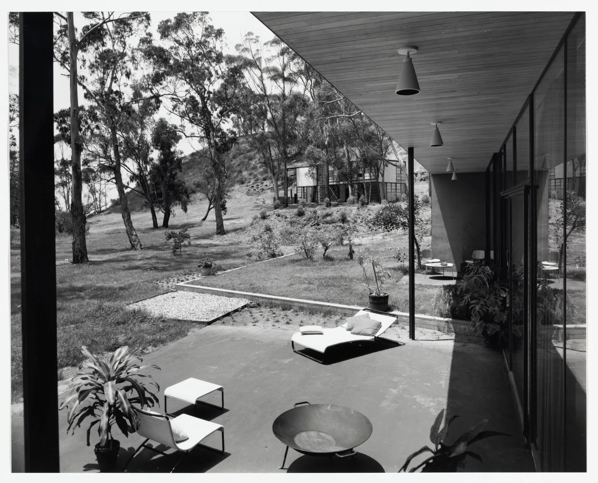 A black and white photo of a minimalist mid-century modern patio featuring a wood plank overhang, chaise lounge, fire pit, and glass sliding doors.
