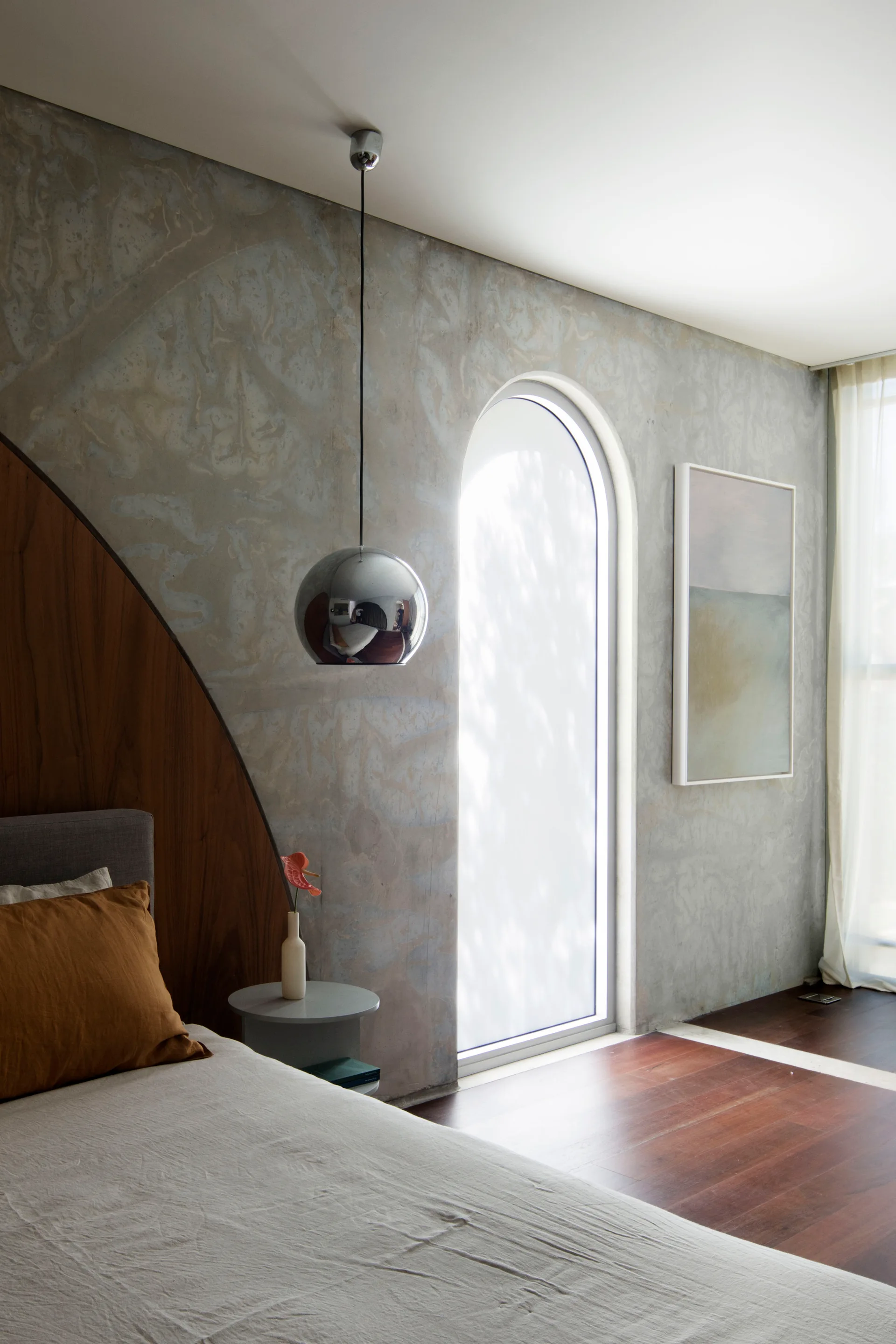 A modern bedroom features a curved walnut wood headboard panel, a textured plaster wall, an arched window, and a hanging chrome pendant lamp.