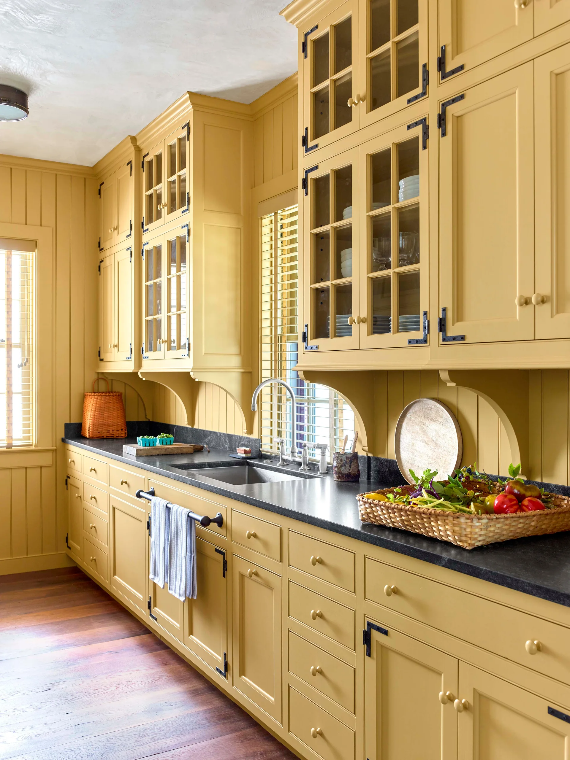 A kitchen features mustard yellow cabinets, dark stone countertops, vertical beadboard walls, and a stainless steel undermount sink.