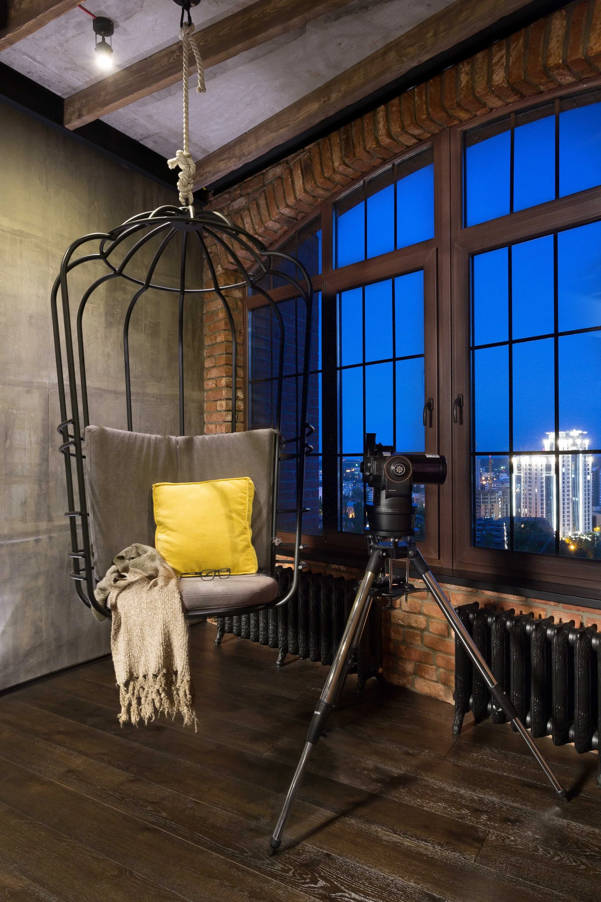 A black metal hanging cage chair with a yellow pillow sits next to a telescope pointing out a large window overlooking a city at night.