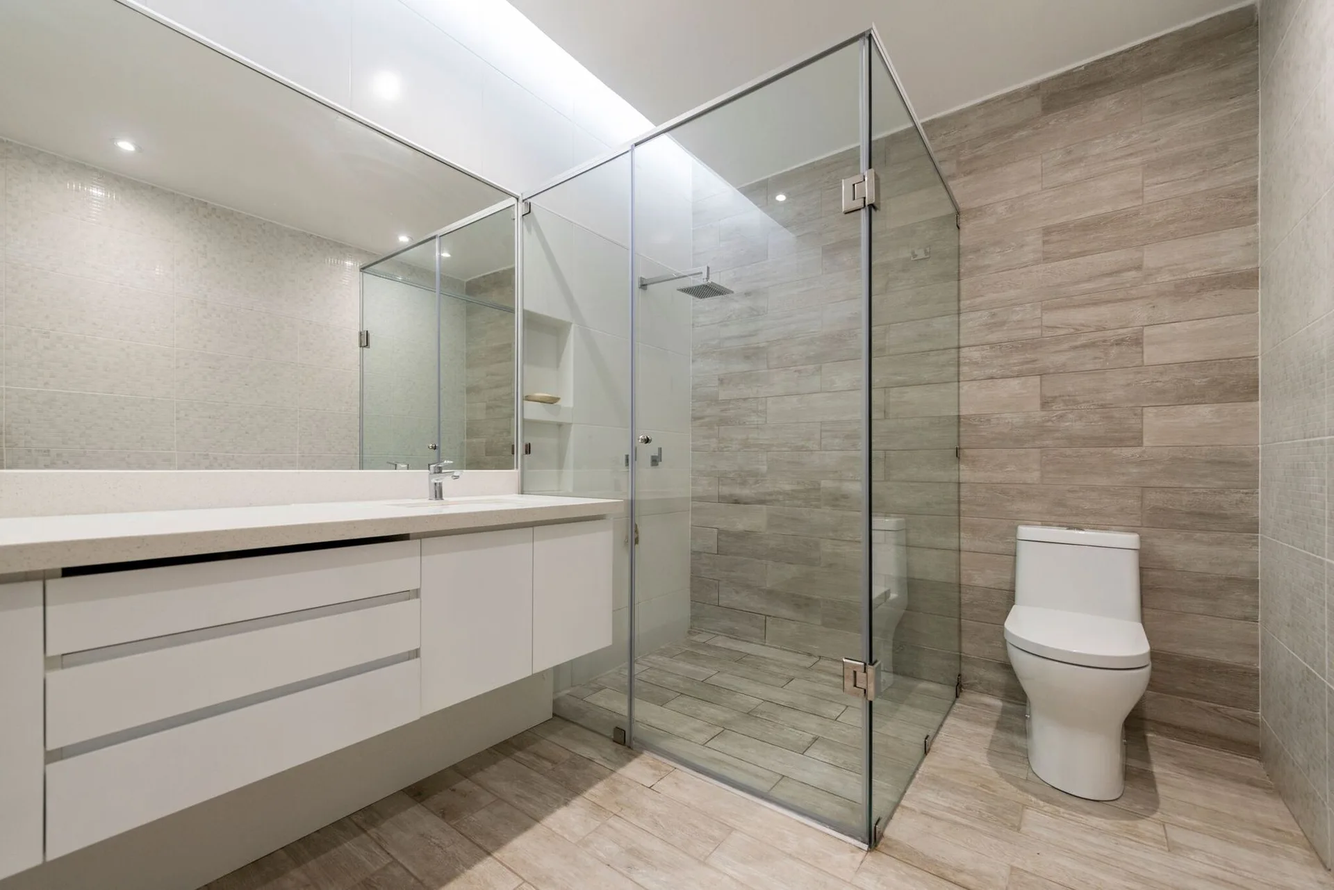 A modern bathroom featuring a white floating vanity, a glass-enclosed shower, a toilet, and wood-look tiled floors and walls.