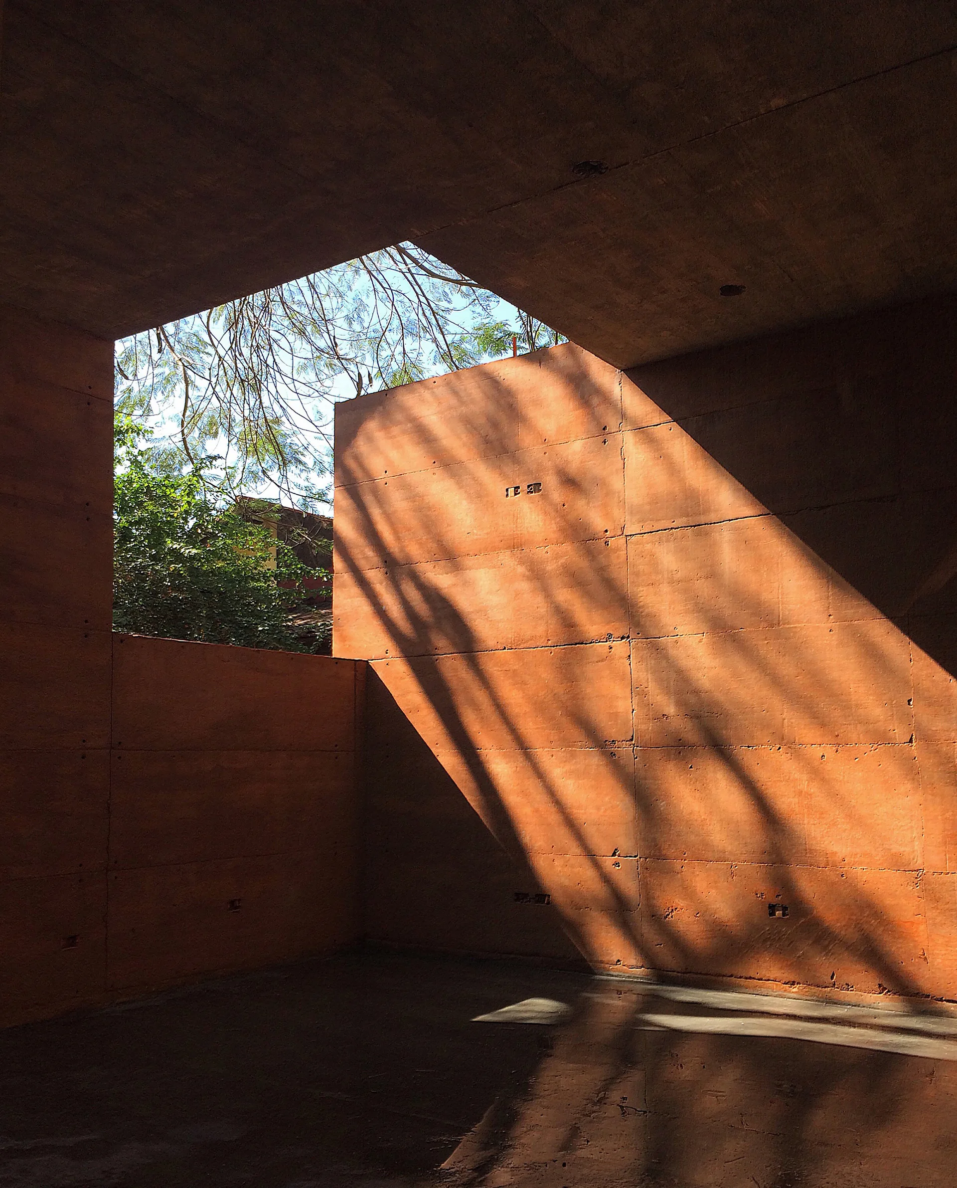 A minimalist room with terracotta-colored concrete panels and a large geometric skylight casting dramatic tree branch shadows on the walls.