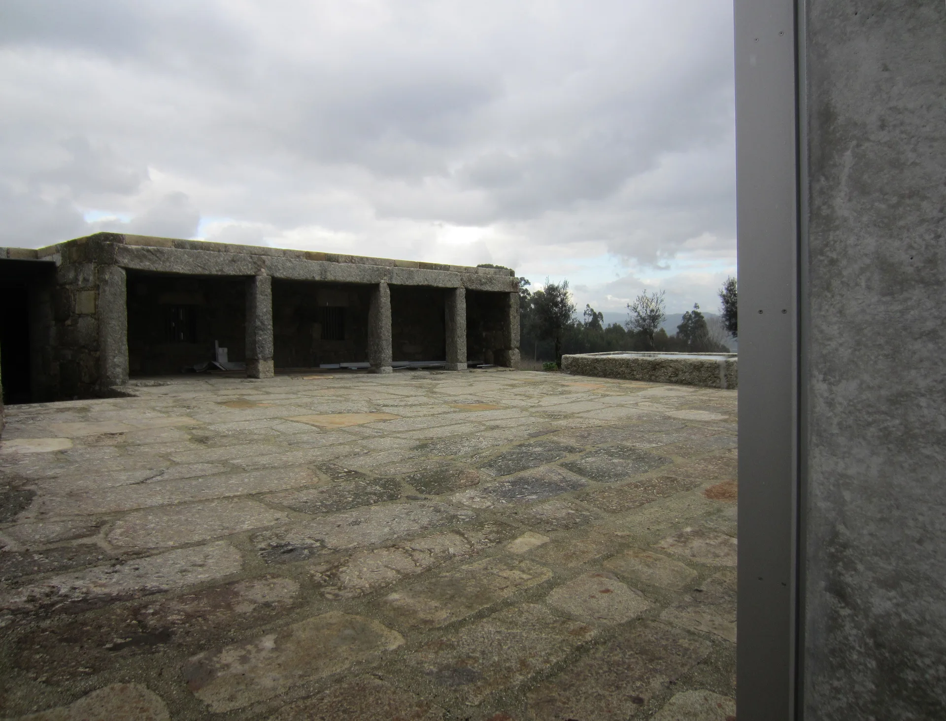 A wide outdoor rustic flagstone patio features a heavy stone covered pavilion supported by thick pillars under an overcast sky.
