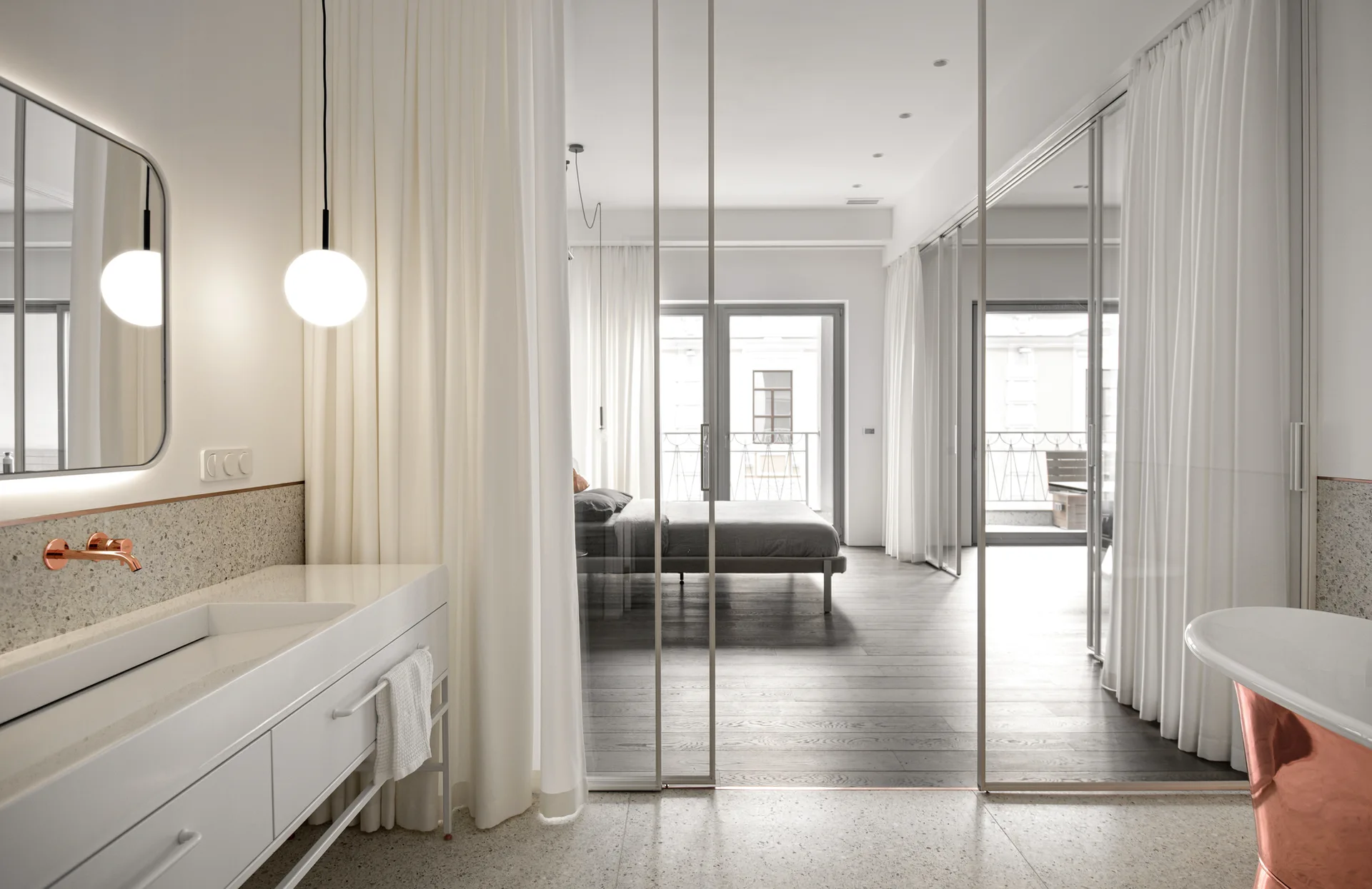 A minimalist bathroom with a white vanity and copper bathtub separated from a bedroom by tall sliding glass doors.