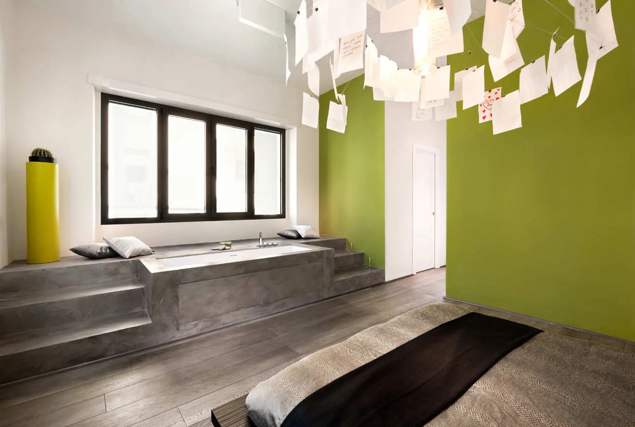 A modern bedroom features a sunken bathtub integrated into a grey concrete platform next to a lime green accent wall and a window.