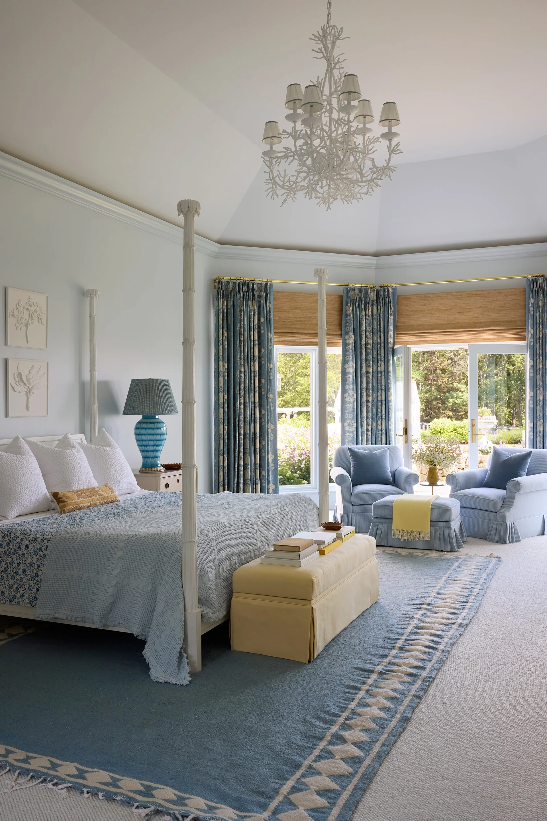 A light blue bedroom features a white four-poster bed, a yellow footboard bench, and blue armchairs by French doors.