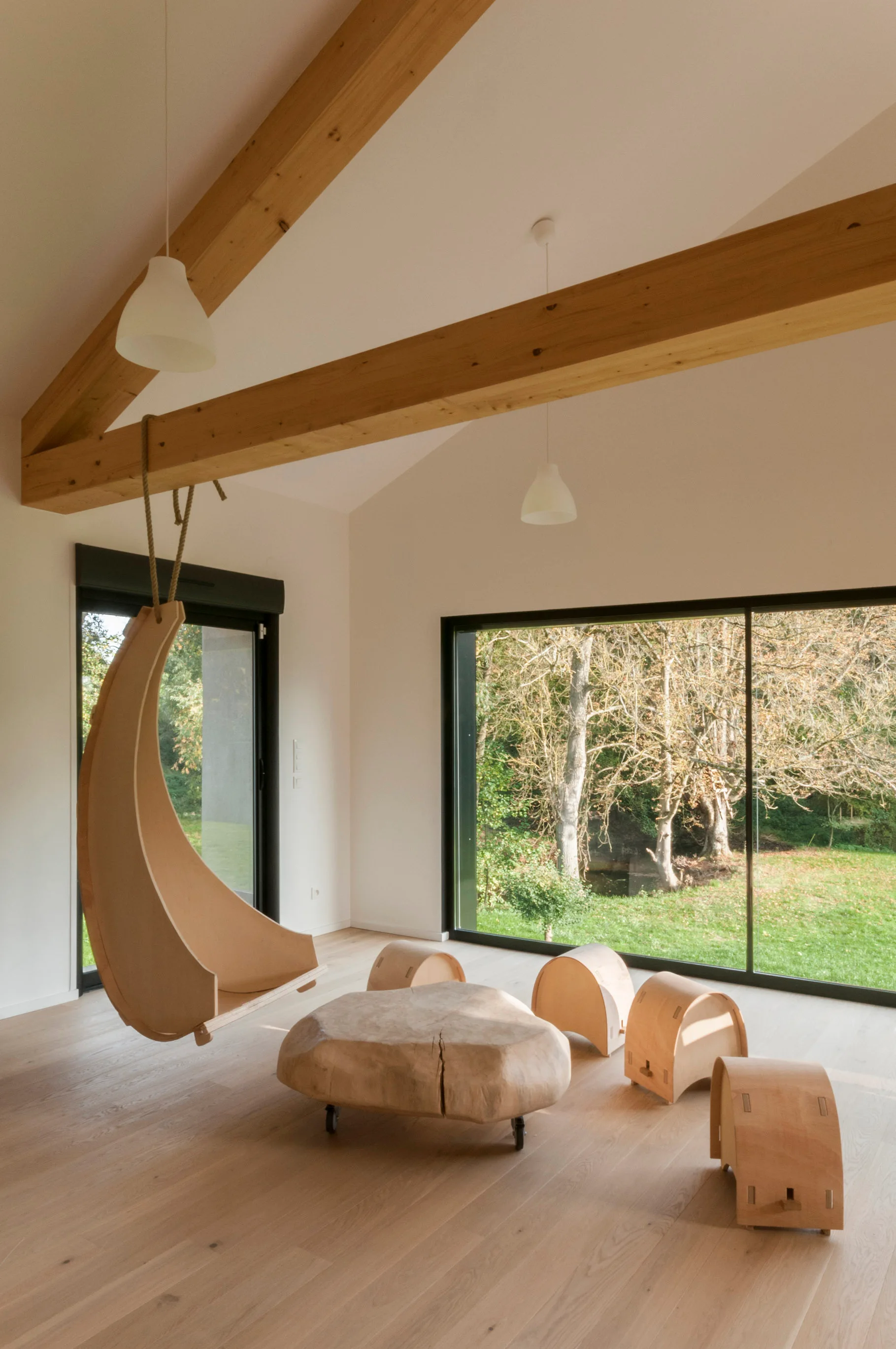 A minimalist living room with white walls, exposed wood ceiling beams, a crescent-shaped hanging chair, a live-edge coffee table, and large windows.