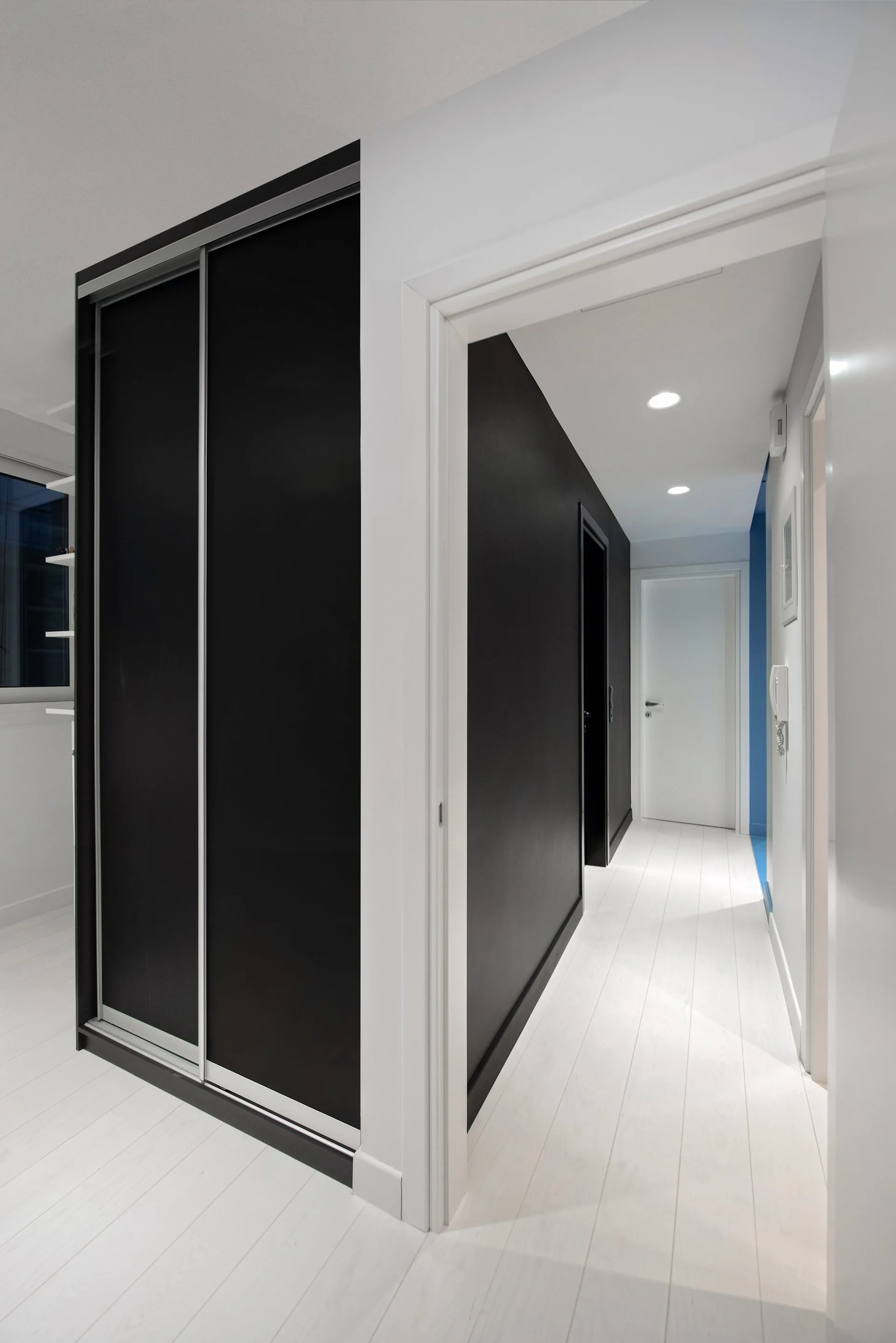 A minimalist hallway features white plank floors, stark white walls, and a large built-in wardrobe with matte black sliding doors.