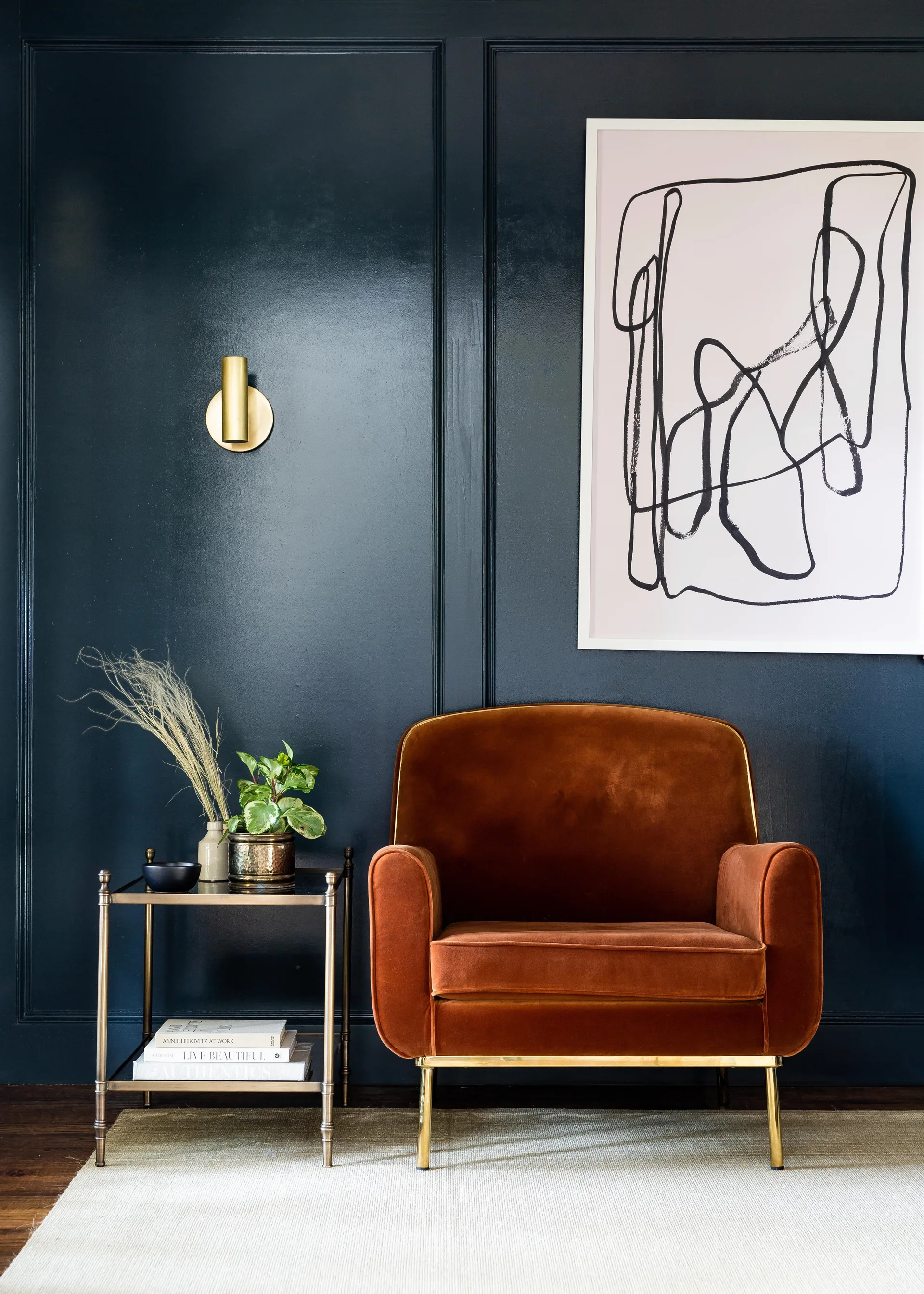 A rust velvet armchair sits next to a brass side table against a dark navy paneled wall decorated with abstract line art and a brass sconce.