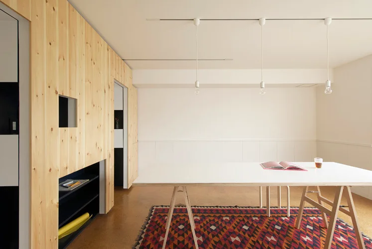 A white trestle table sits on a geometric patterned rug next to a light pine paneled wall with a black recessed shelf.