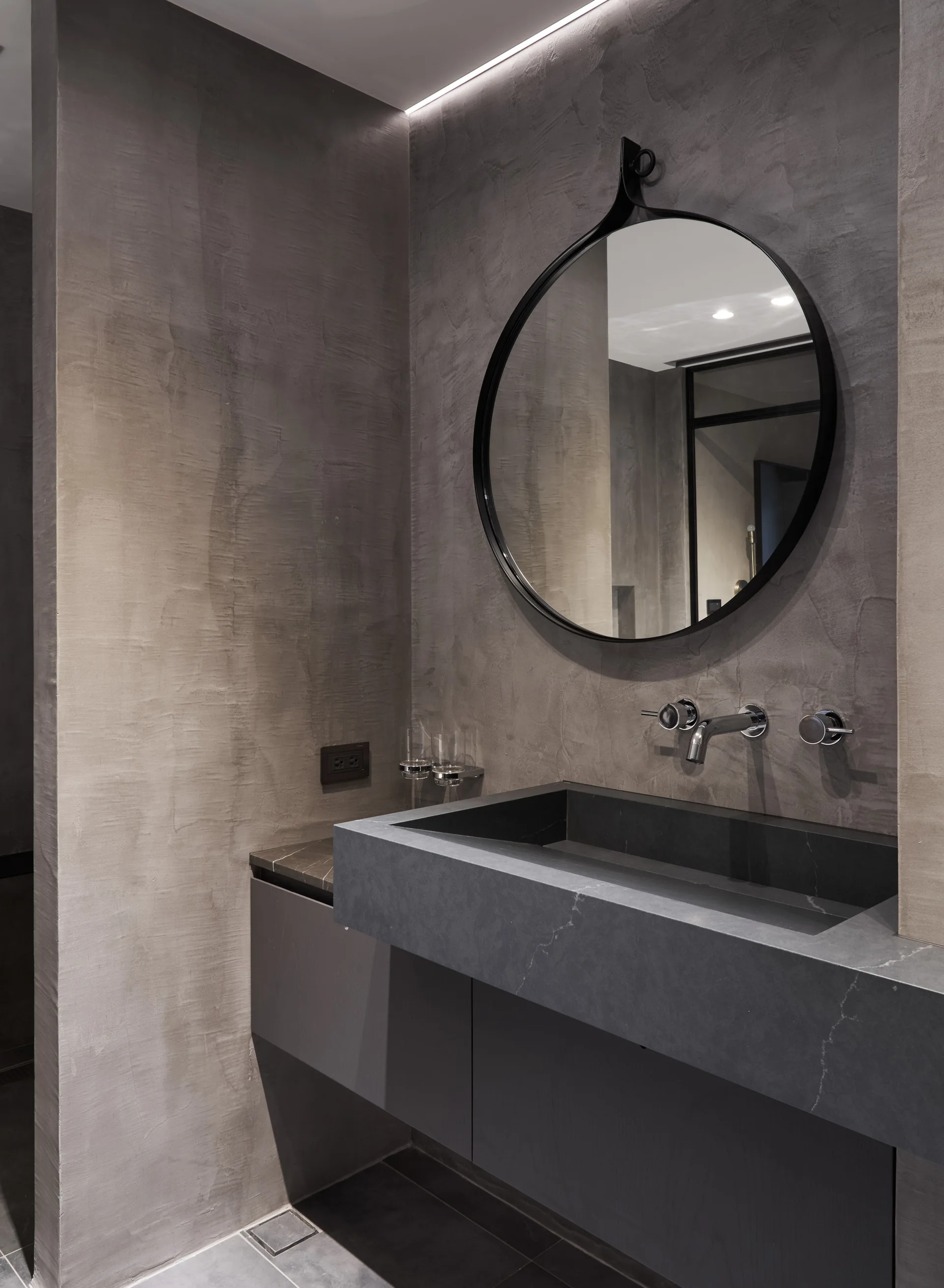 A modern grey bathroom vanity with a dark stone basin faucet wall-mounted under a circular black-framed mirror on a textured plaster wall.