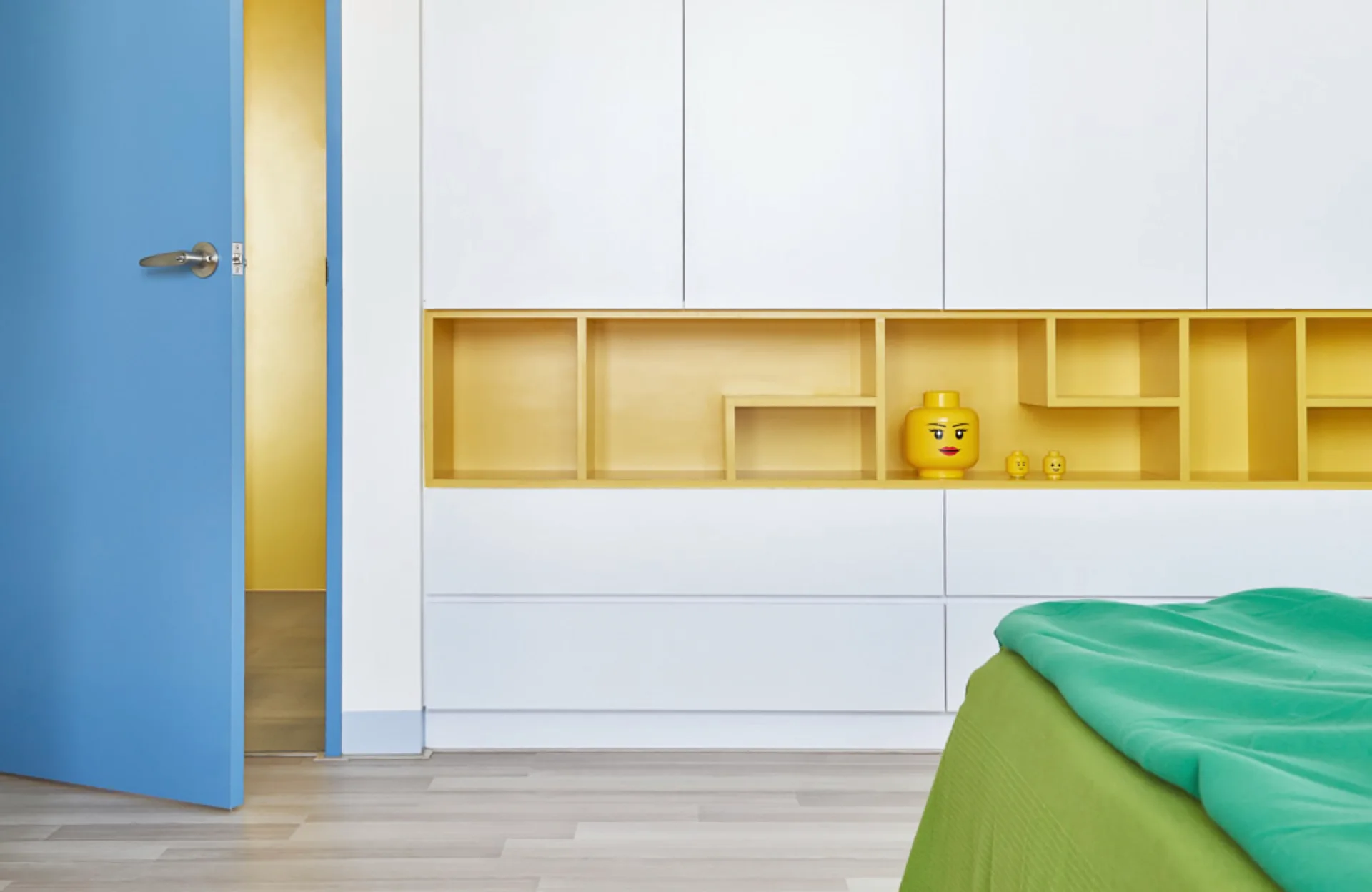A modern kids' room featuring white built-in storage cabinets with a recessed yellow shelf, an open blue door, and a green bed.