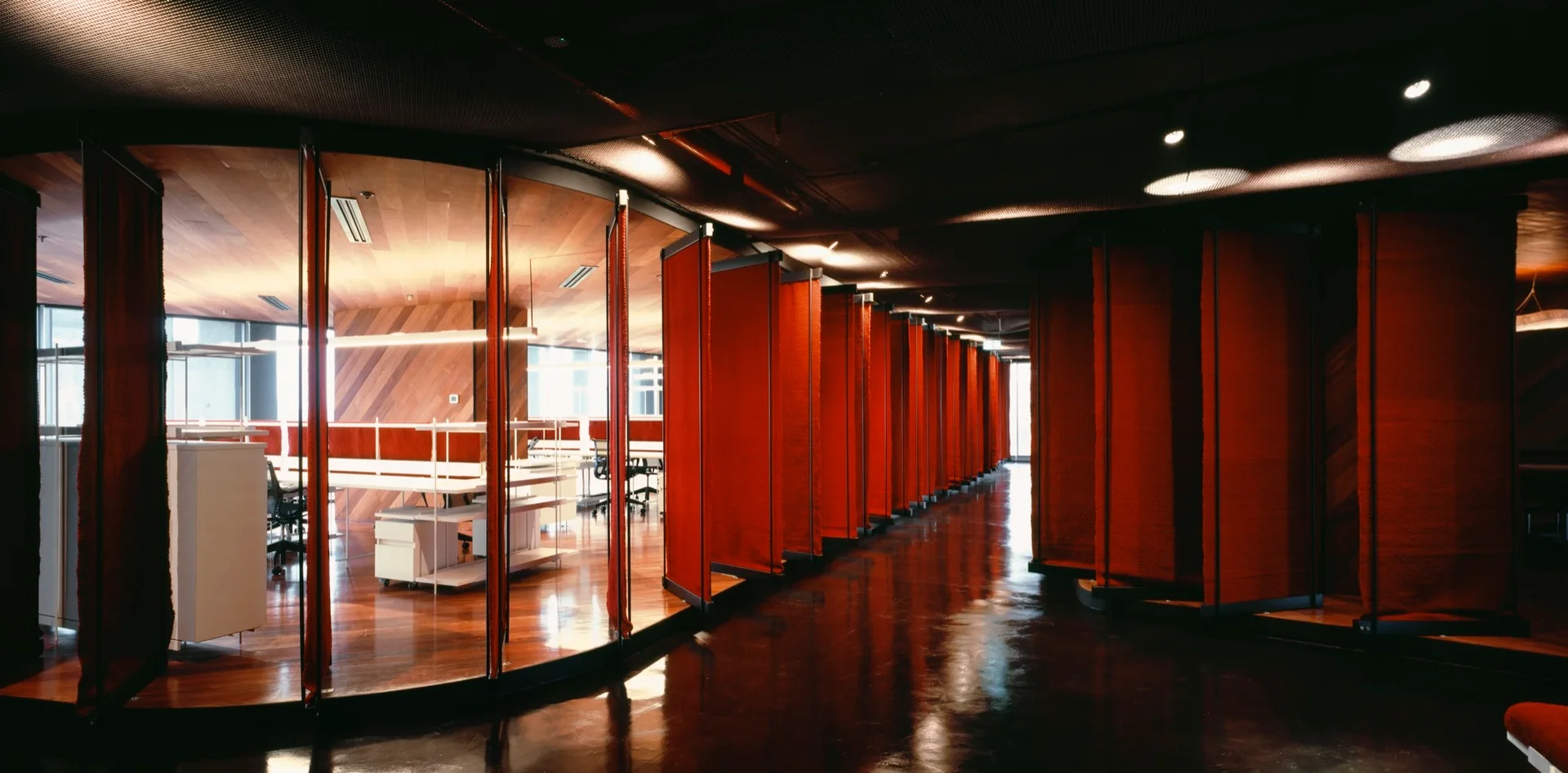 A modern open office space with polished wood floors and a long row of suspended red fabric partition panels separating workstations.