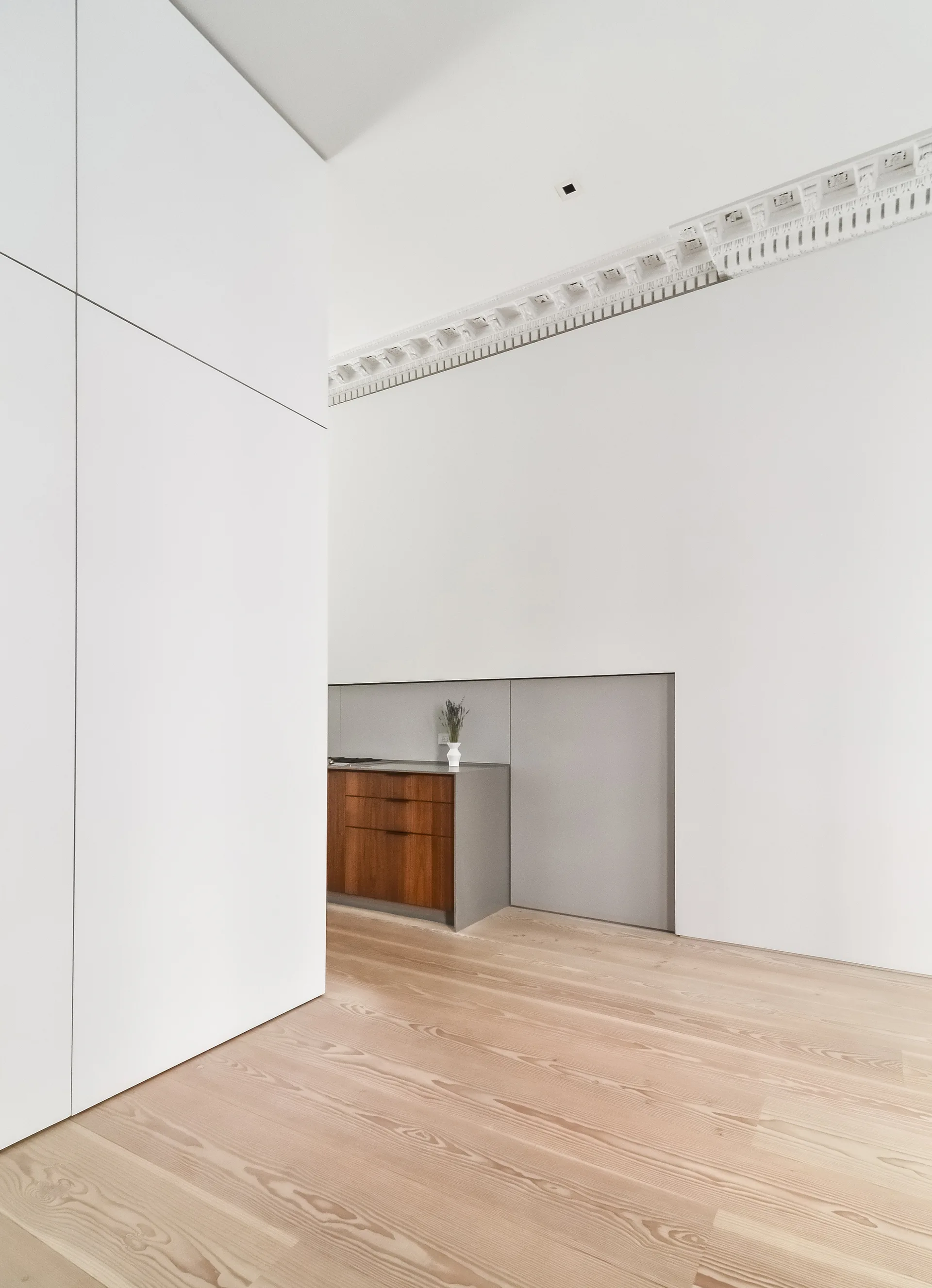 A minimalist kitchen nook with wood base cabinets and a grey countertop, set against flat white walls and ornate ceiling moulding.