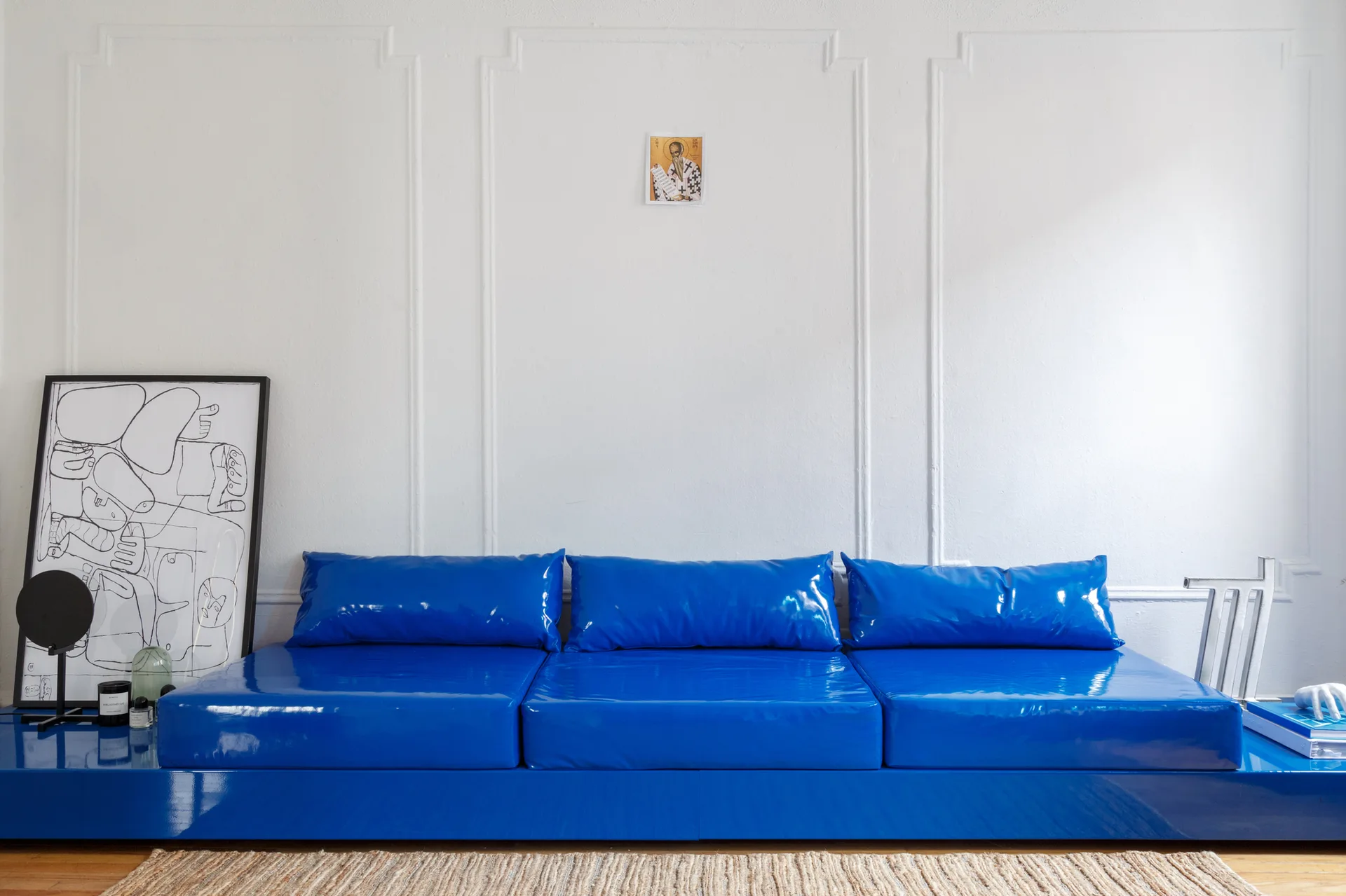A shiny, high-gloss blue vinyl sofa is positioned against a white wall decorated with traditional molding panels, next to leaning framed art.