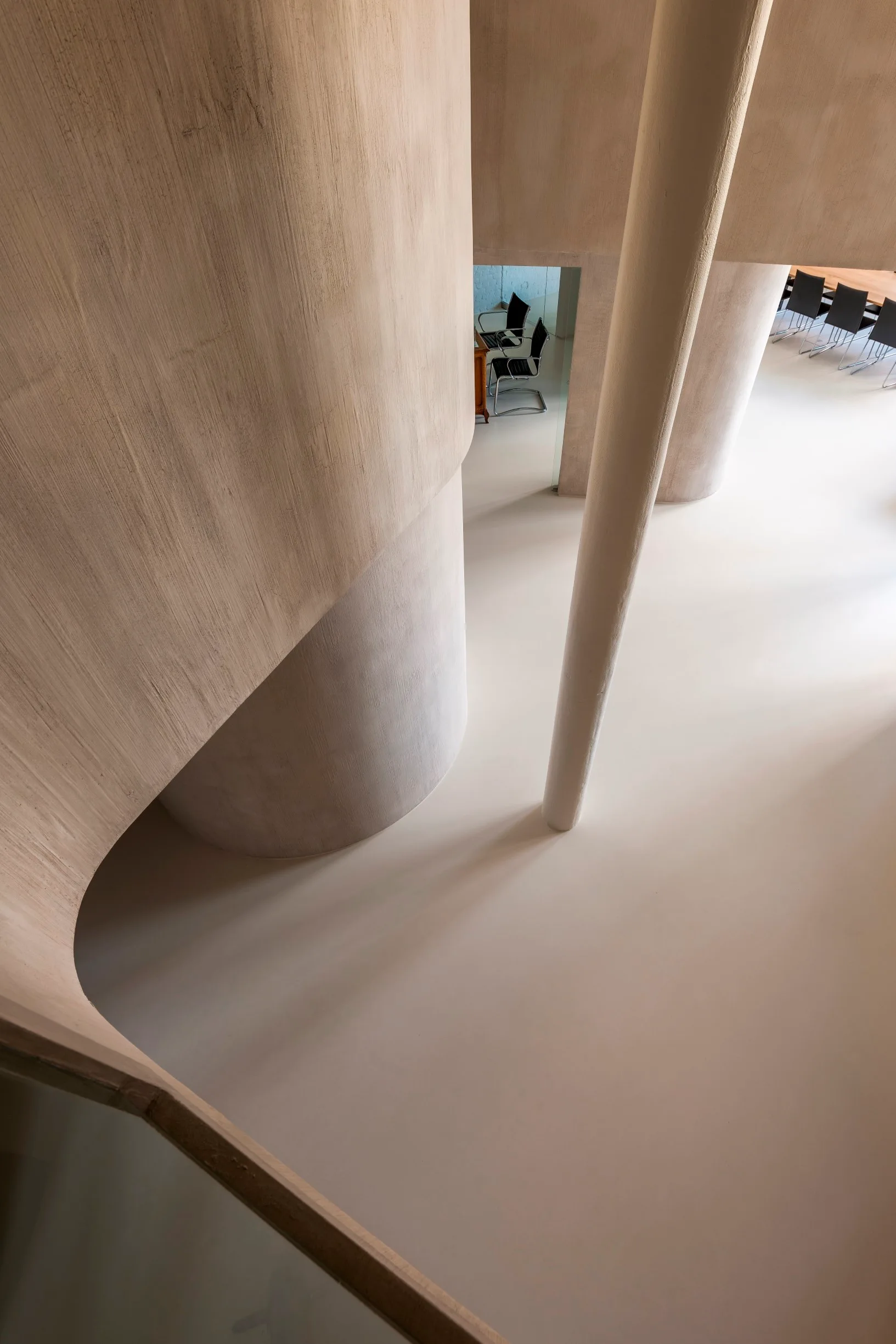 High angle view of a curved plaster staircase with textured beige walls, a slender column, and smooth seamless flooring in a minimalist interior.
