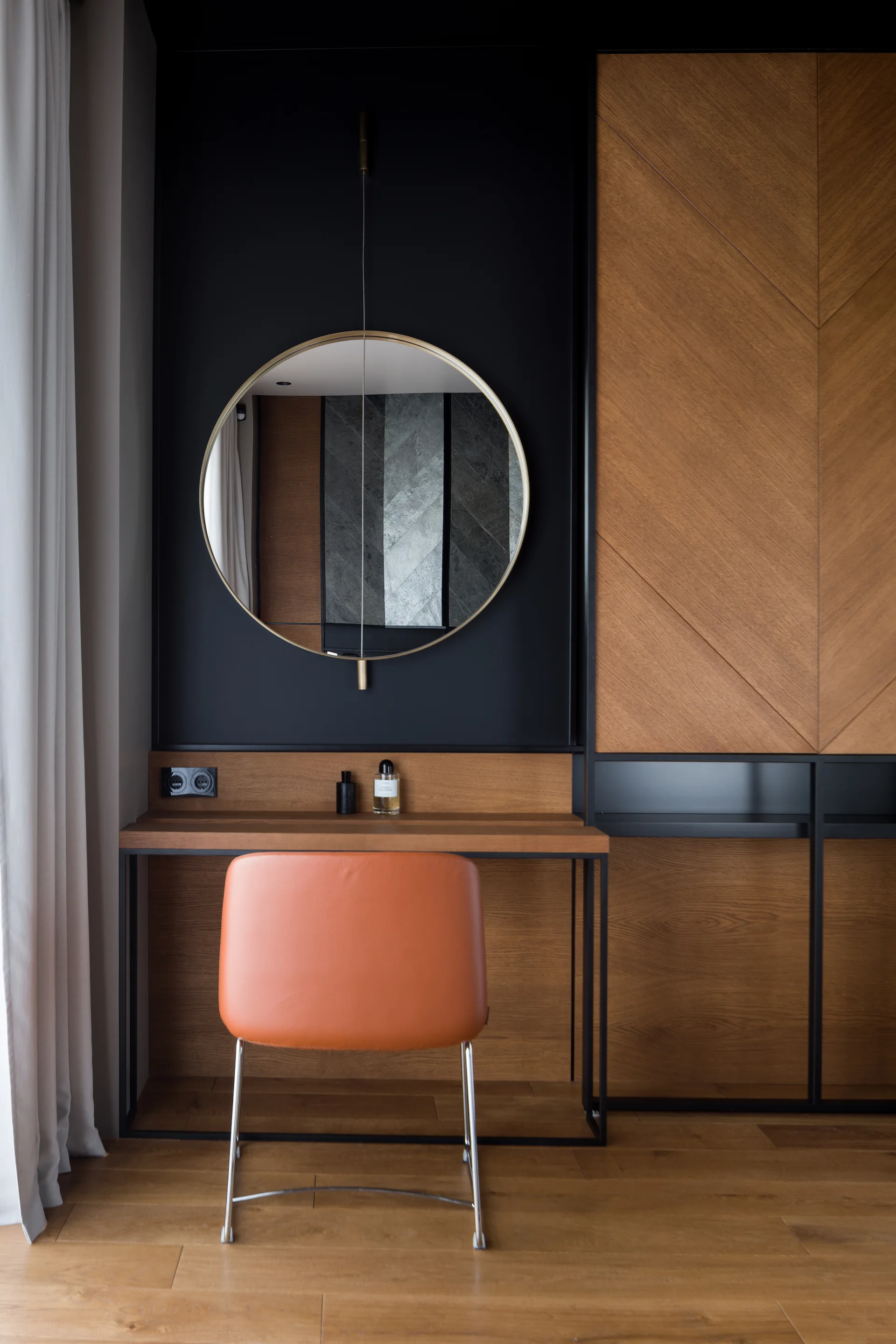 A modern built-in wooden vanity with a round mirror against a matte black wall panel and an orange leather-like chair.