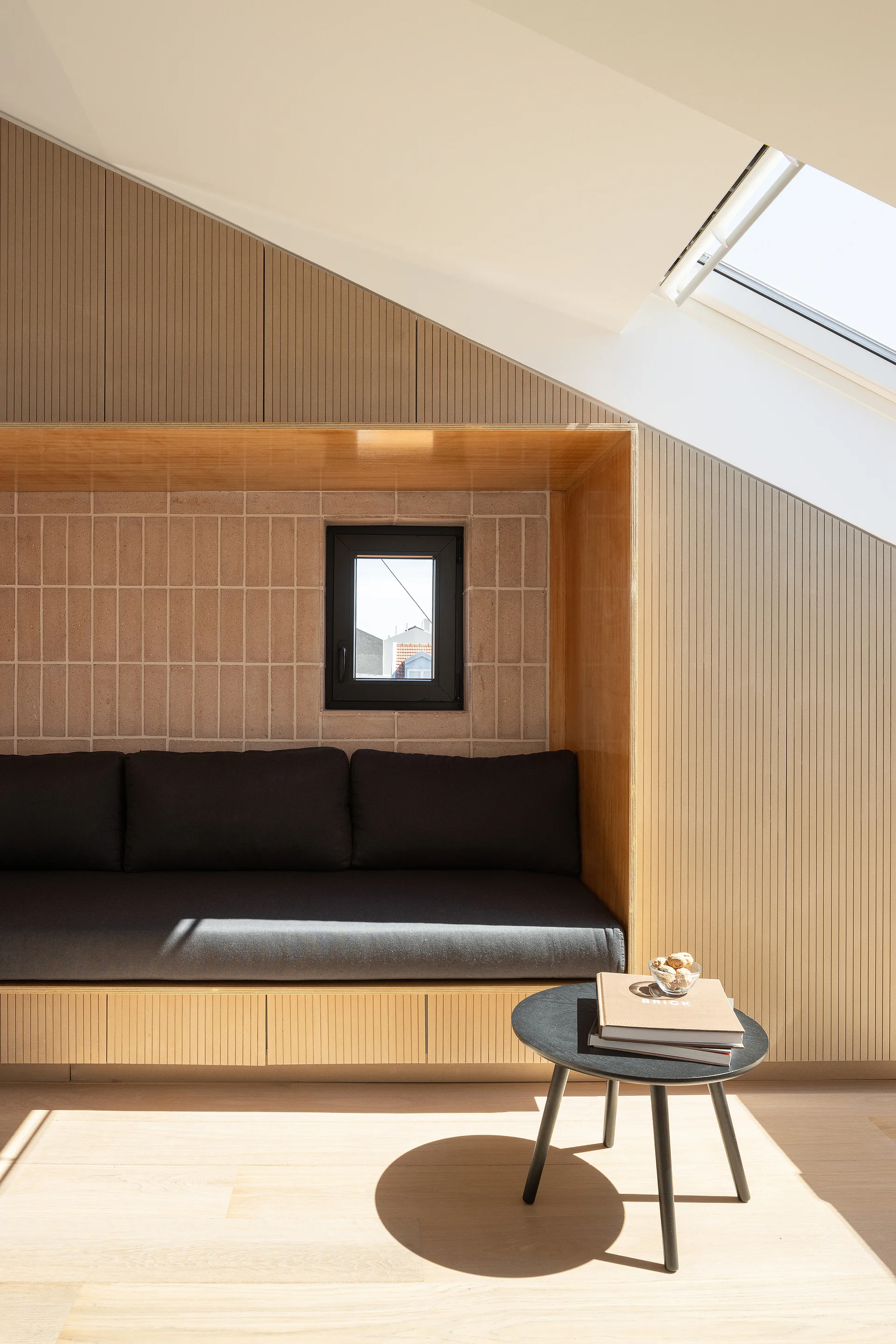 A custom wood-framed built-in bench with grey cushions sits beneath a sloped ceiling and skylight next to a small black coffee table.