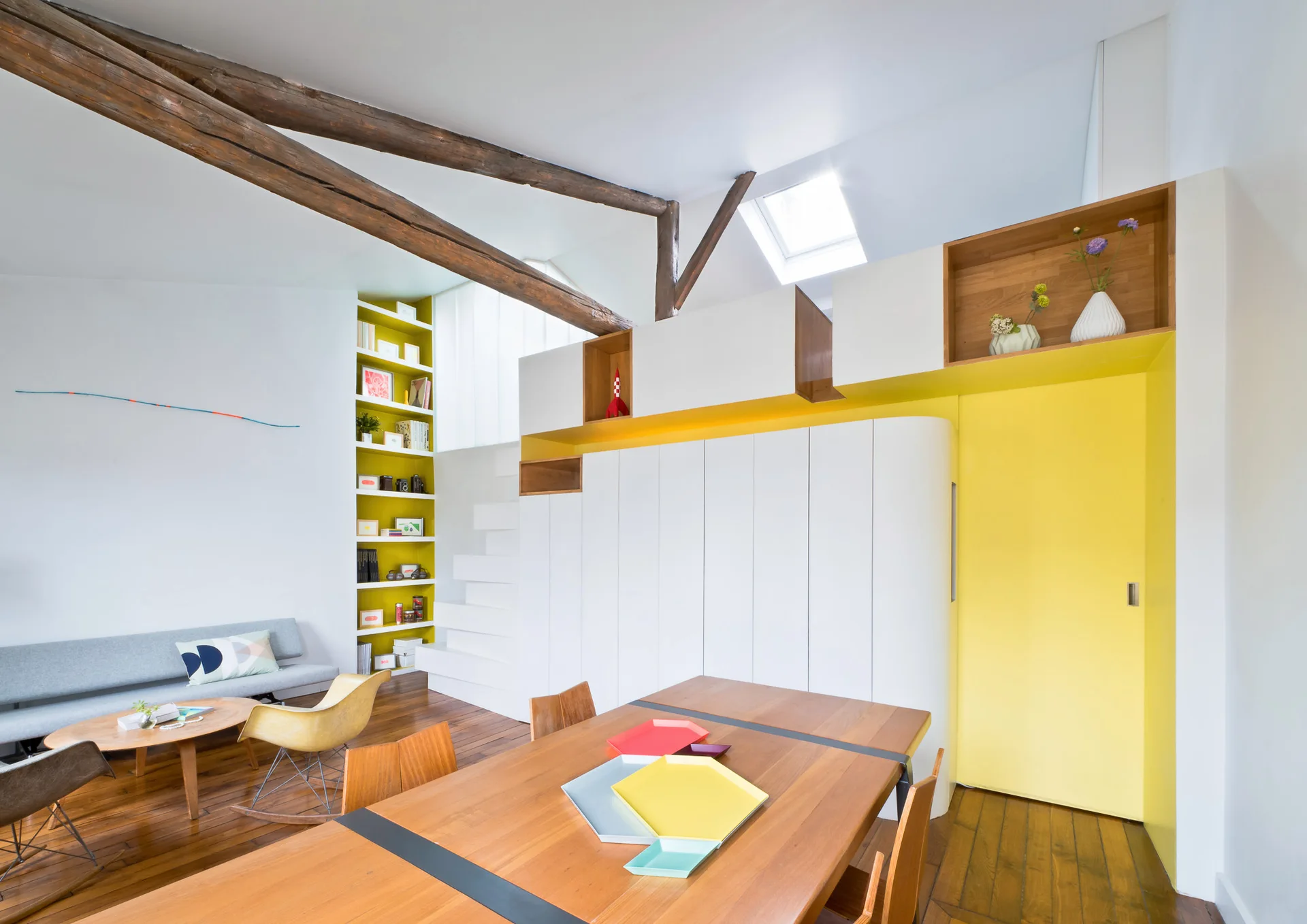 An open living and dining area featuring custom white storage cabinets with yellow accents, dark exposed ceiling beams, and a wood dining table.