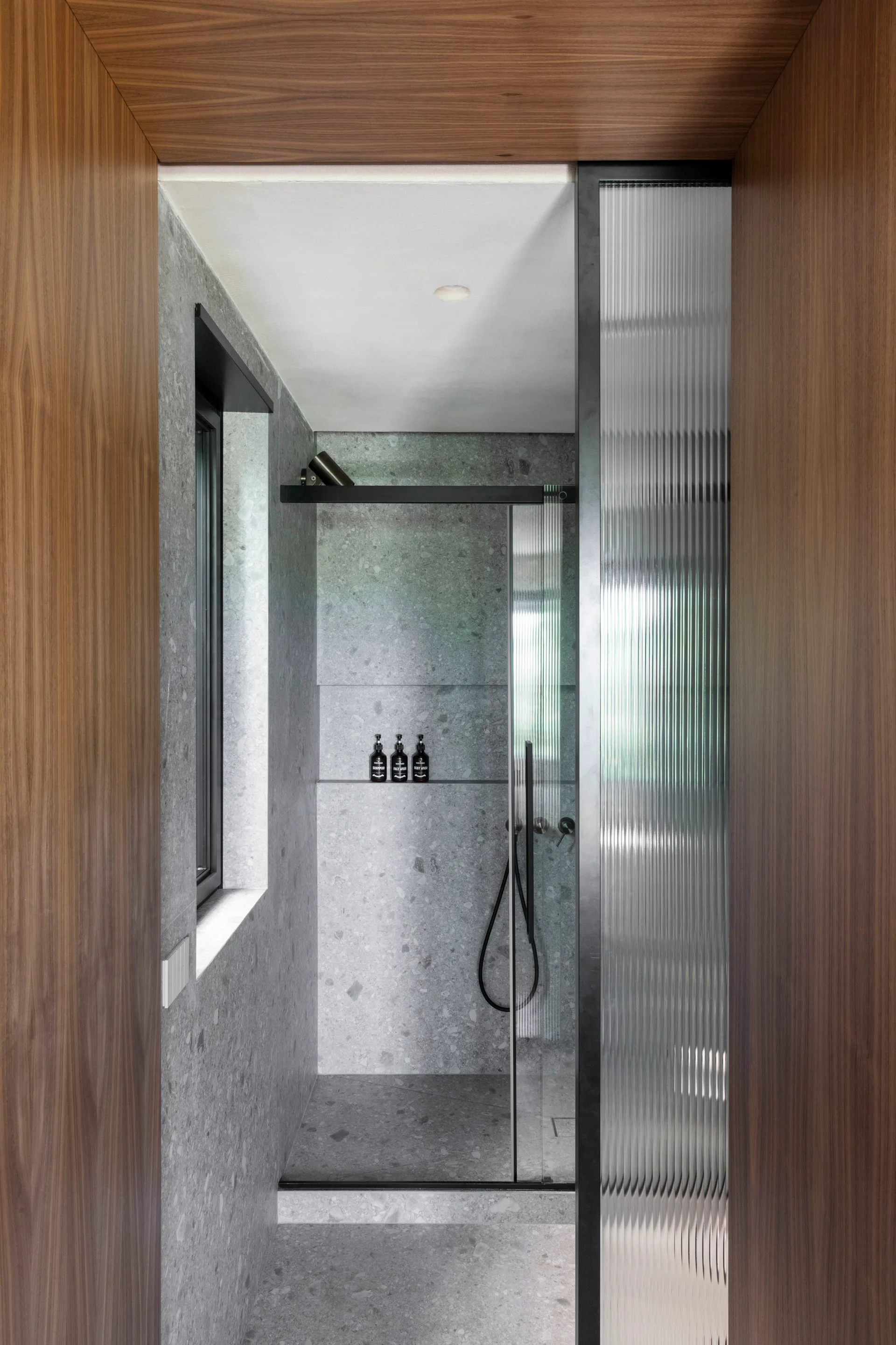 A walk-in shower with grey stone tile walls is viewed through a wood-paneled threshold next to a fluted glass partition.