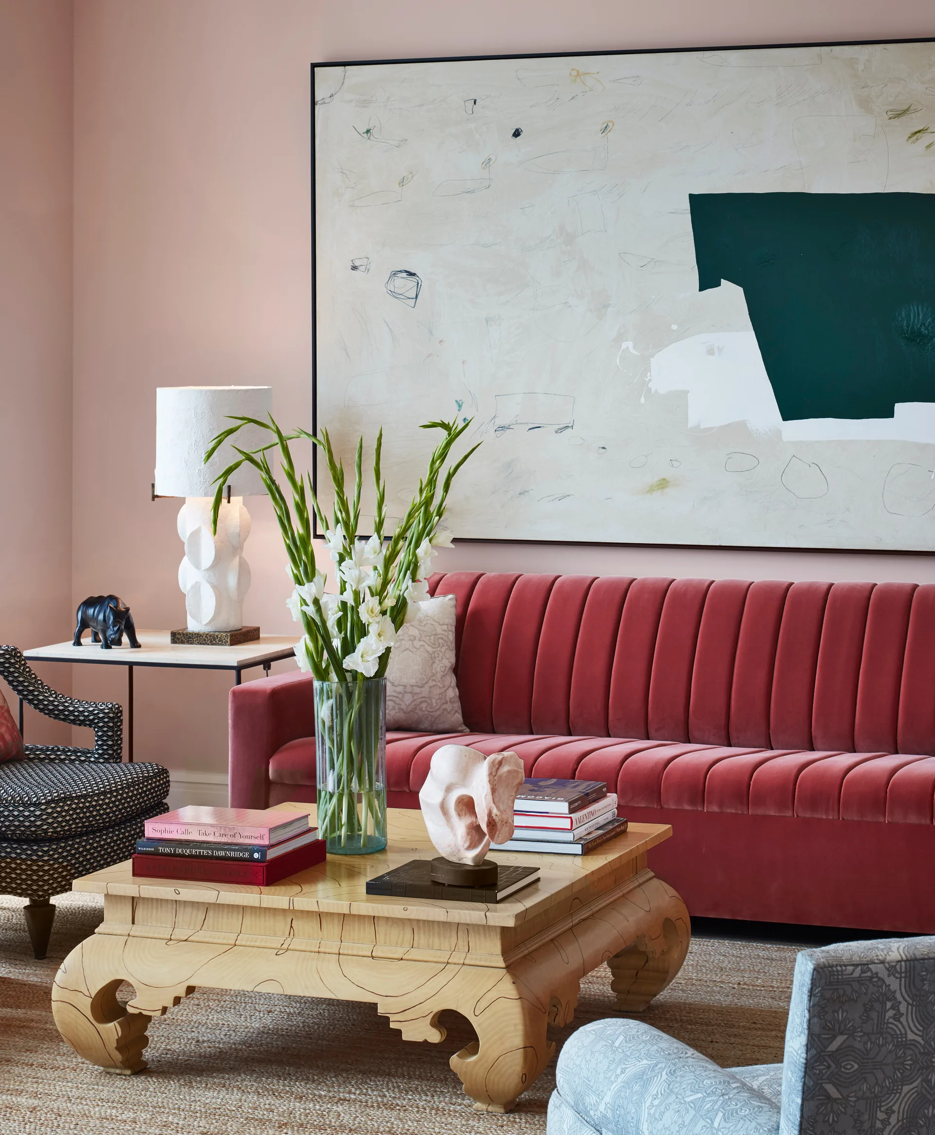 A living room featuring a channel-tufted red velvet sofa, a carved wood coffee table with books, and a large abstract painting on a pink wall.