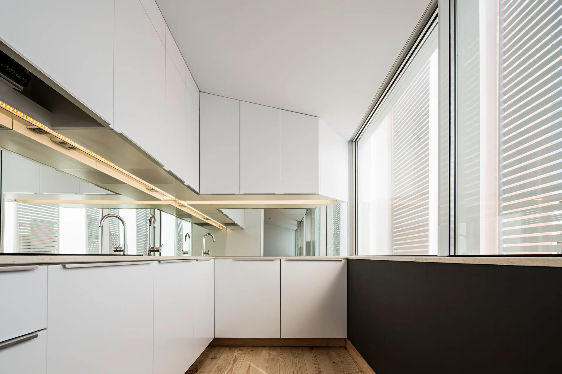 A minimalist white kitchen features flat-panel handleless cabinets, a mirrored backsplash, and light wood plank flooring.