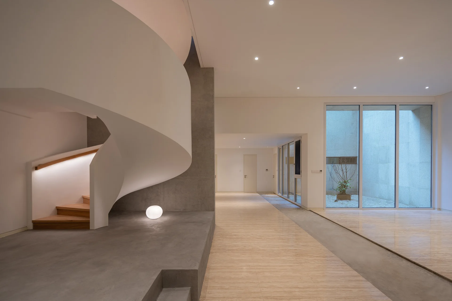 A minimalist foyer features a sculptural white spiral staircase with wooden treads, polished stone flooring, and a glass-walled courtyard in the background.