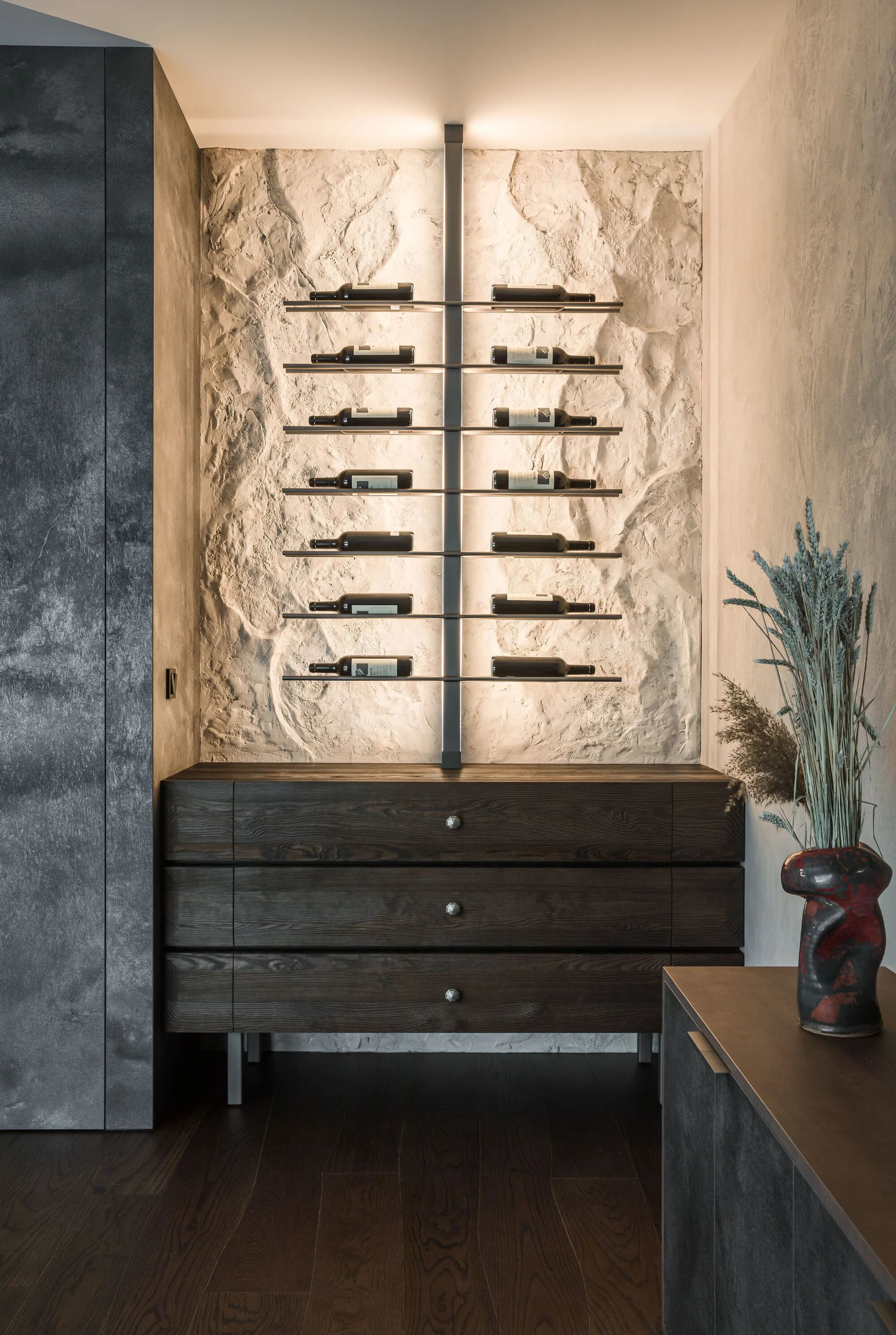 A dark wood cabinet sits beneath a wall-mounted metal wine rack lit by integrated backlighting against a textured stone feature wall.