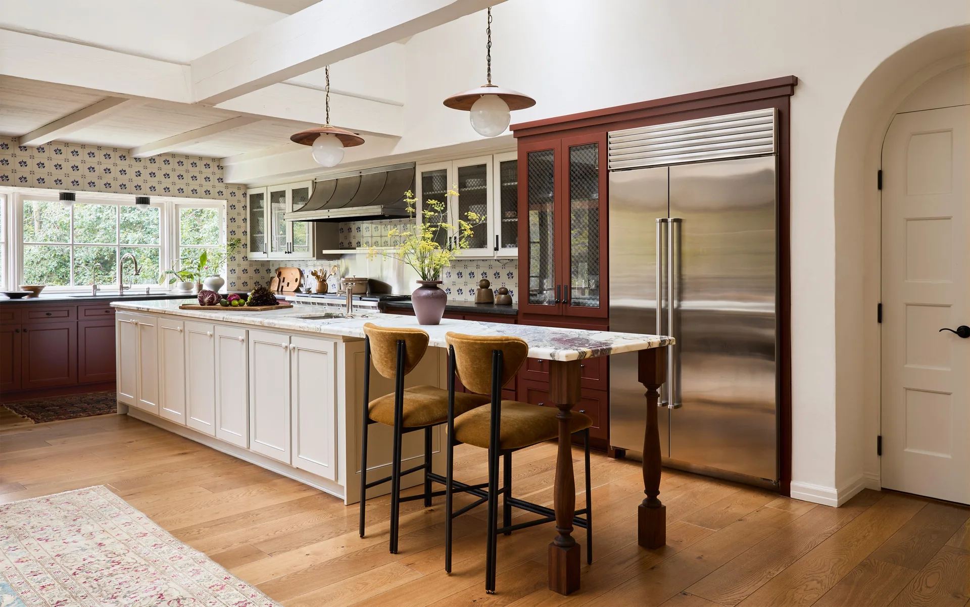 A kitchen featuring a long white island with a marble countertop, mustard yellow bar stools, and deep red cabinetry framing a stainless steel refrigerator.