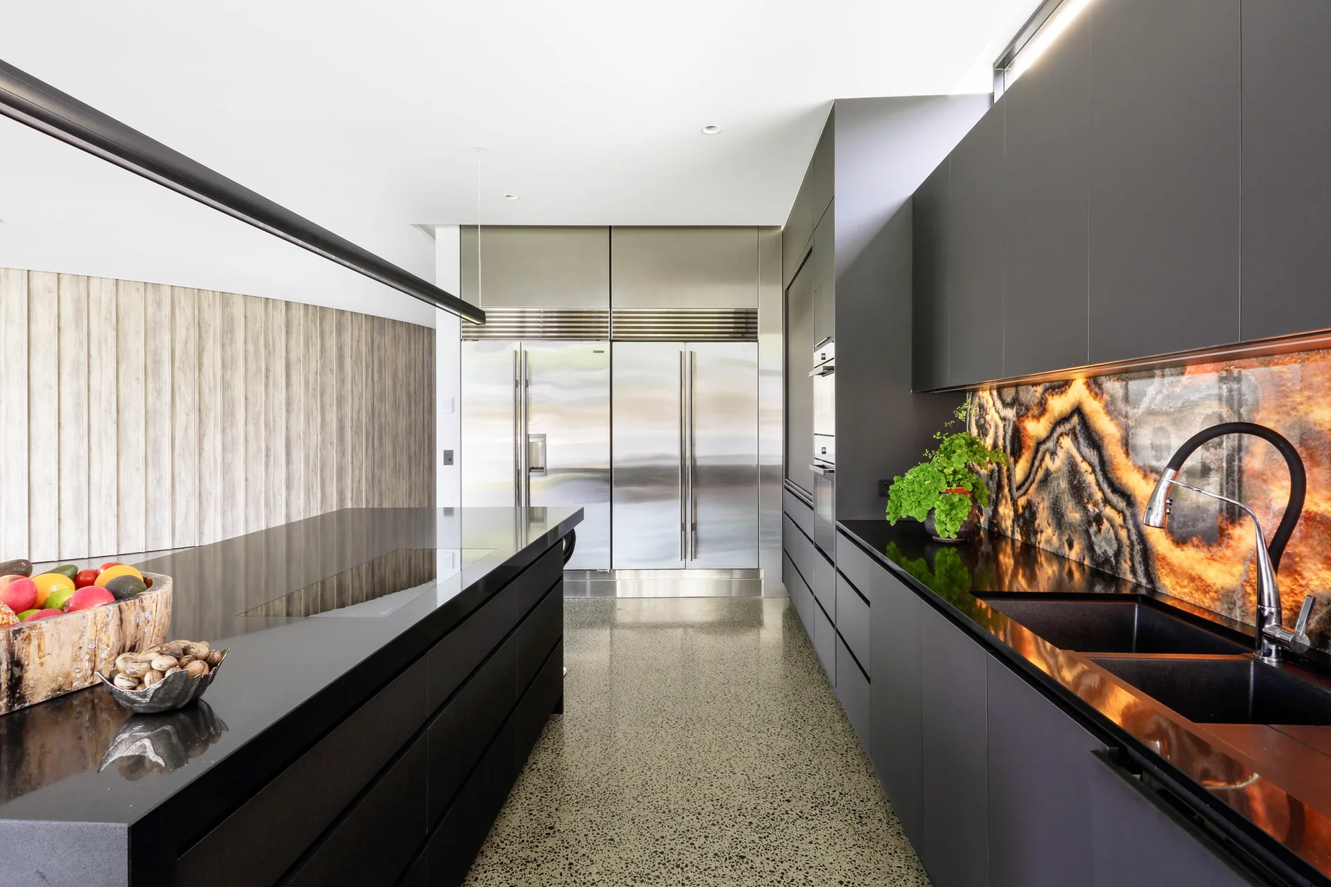 A minimalist kitchen features dark grey matte cabinetry, a black kitchen island, stainless steel refrigerators, and a glowing backlit stone backsplash.